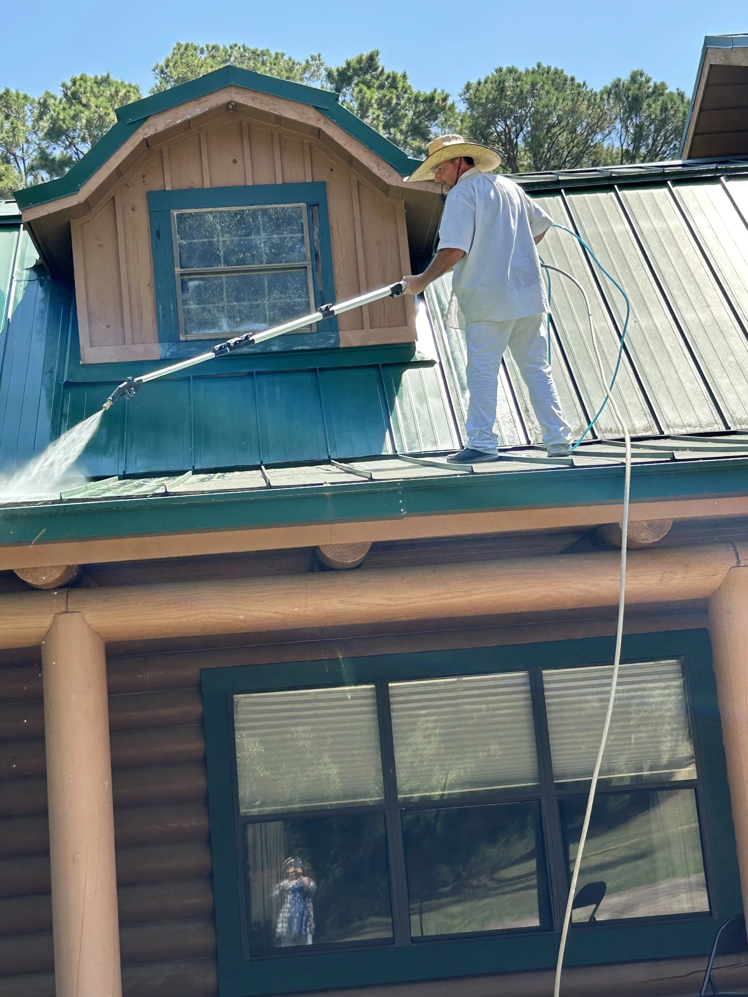 A man wearing a hat and white clothes is pressure washing the roof of a house that has a wooden turret and green metal roof, with trees and a clear sky in the background.