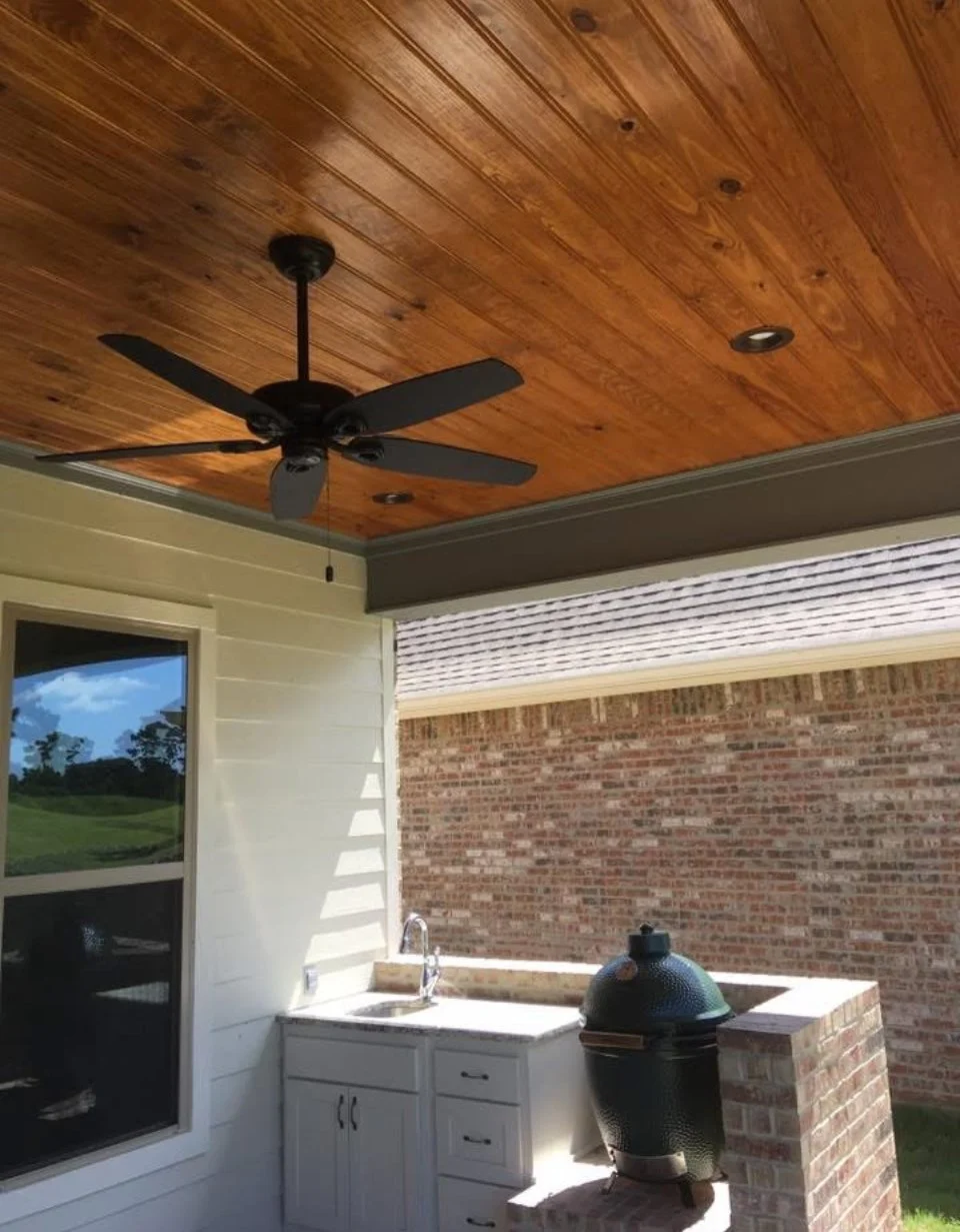 Covered outdoor kitchen area with a black ceiling fan, black grill, white cabinet, sink, and brick wall.