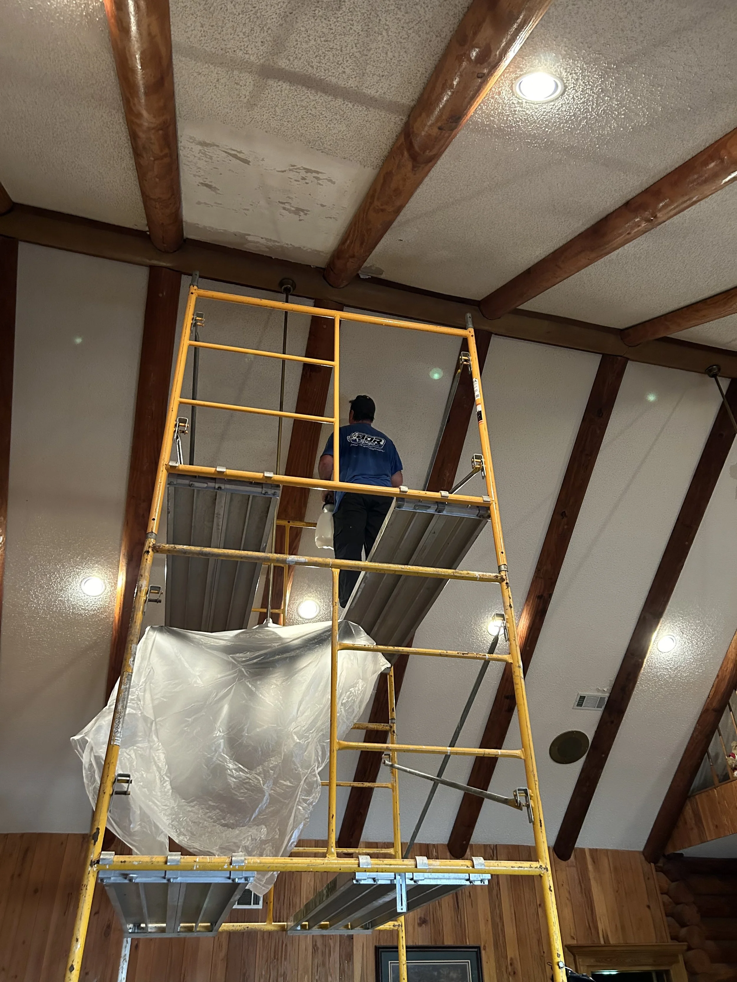 A person standing on a yellow scaffolding measuring or inspecting a ceiling with wood beams, in a room with wood paneled walls and recessed lighting.
