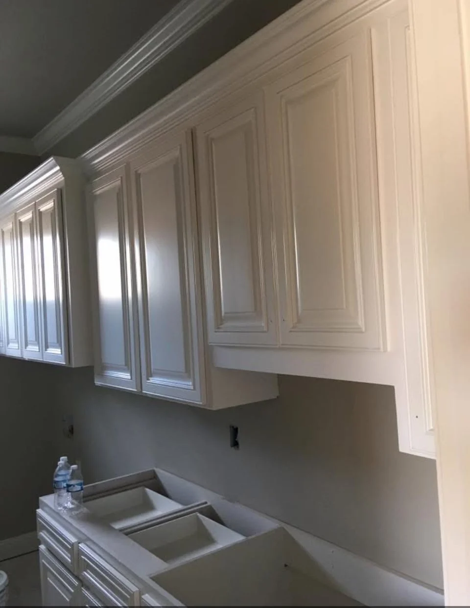 White kitchen cabinets installed, with some upper cabinets and partial lower cabinetry. Two water bottles are on the countertop, and there are no visible appliances or fixtures yet.