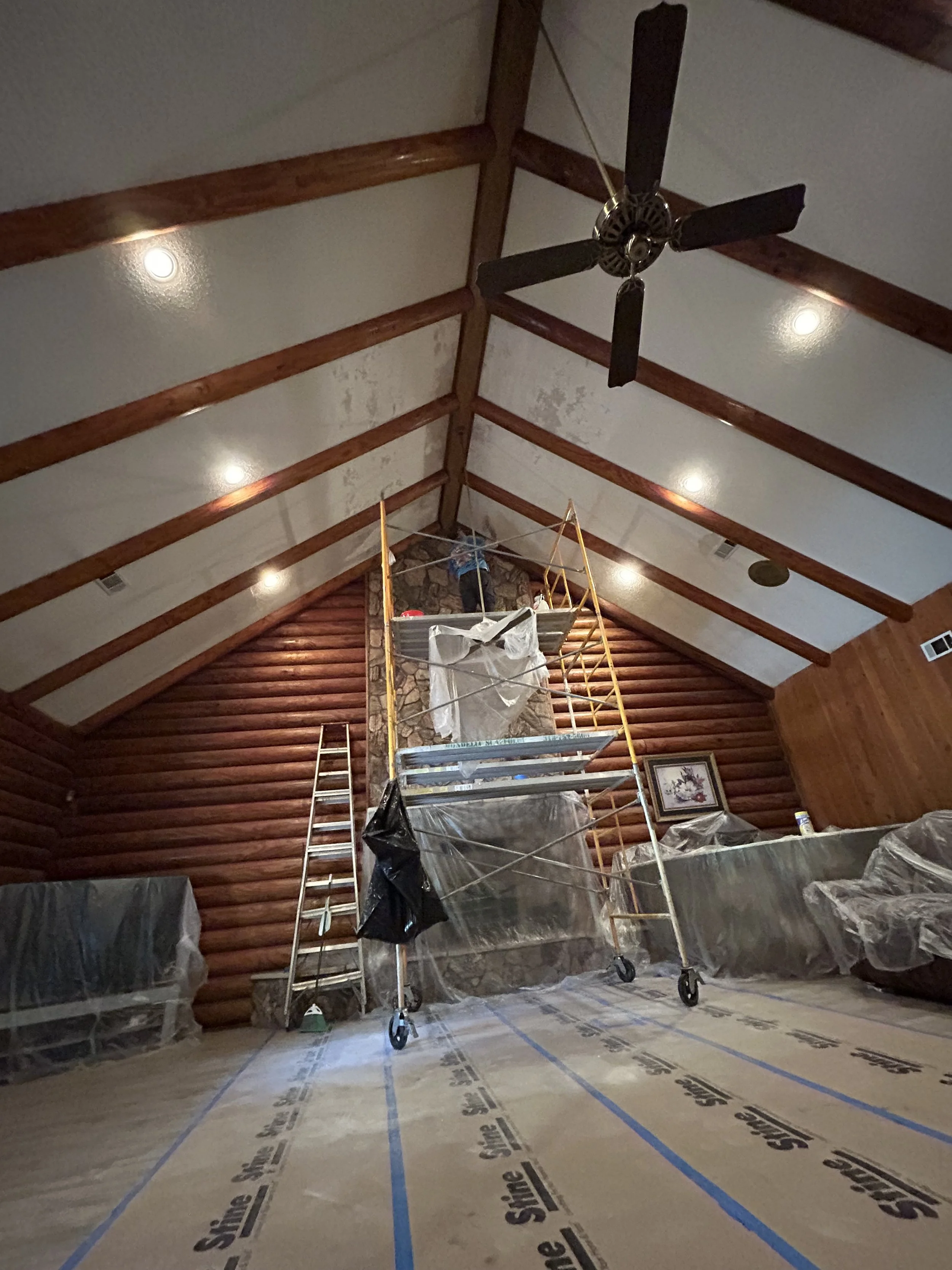 Interior of a log cabin-style room under renovation, with scaffolding, ladders, and furniture covered in plastic, a stone fireplace, and a ceiling fan.