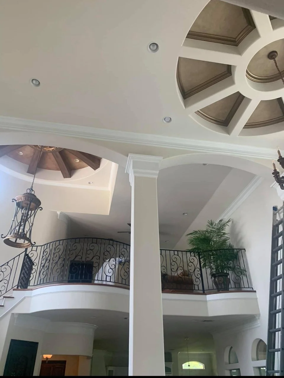 Interior view of a luxurious two-story home with a high ceiling, decorative molding, chandelier, and railing around an upper balcony with potted plants.