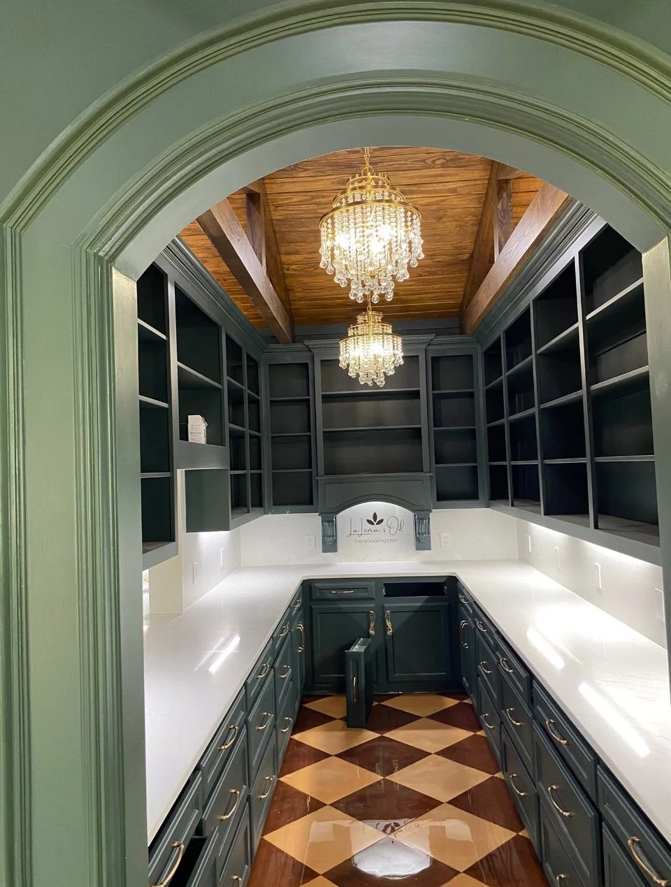 View of a butler's pantry with high dark cabinets, white countertops, and two chandeliers hanging from a wooden ceiling with exposed beams, framed by an arched doorway.