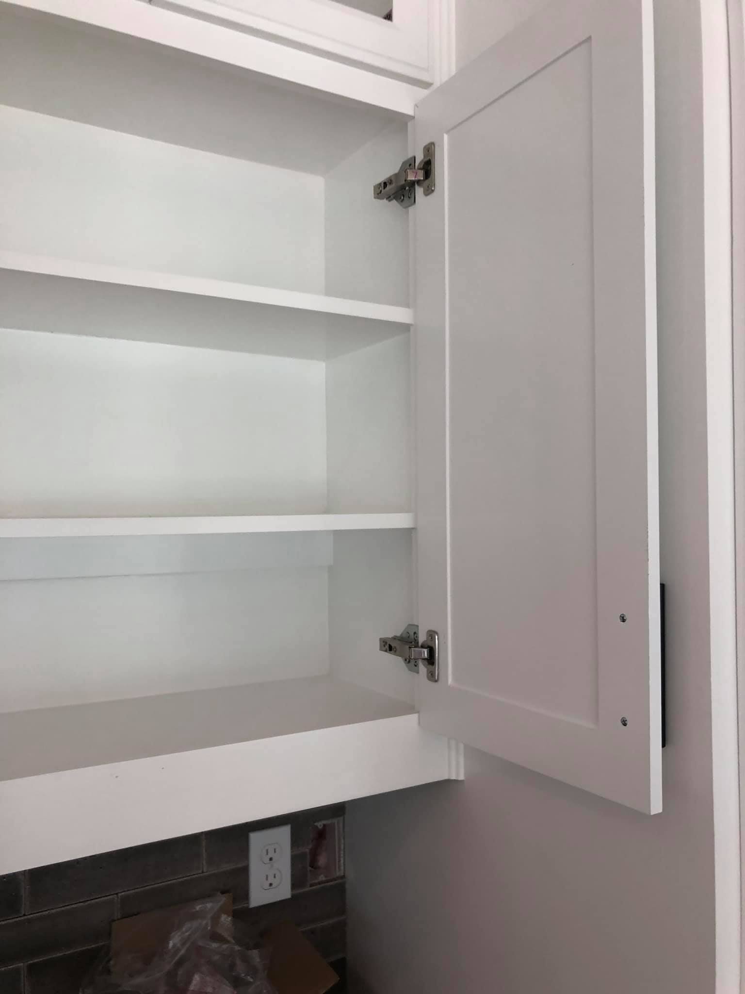 Empty white kitchen cabinet with open door, shelving inside, power outlets below, and a backsplash with dark tiles.