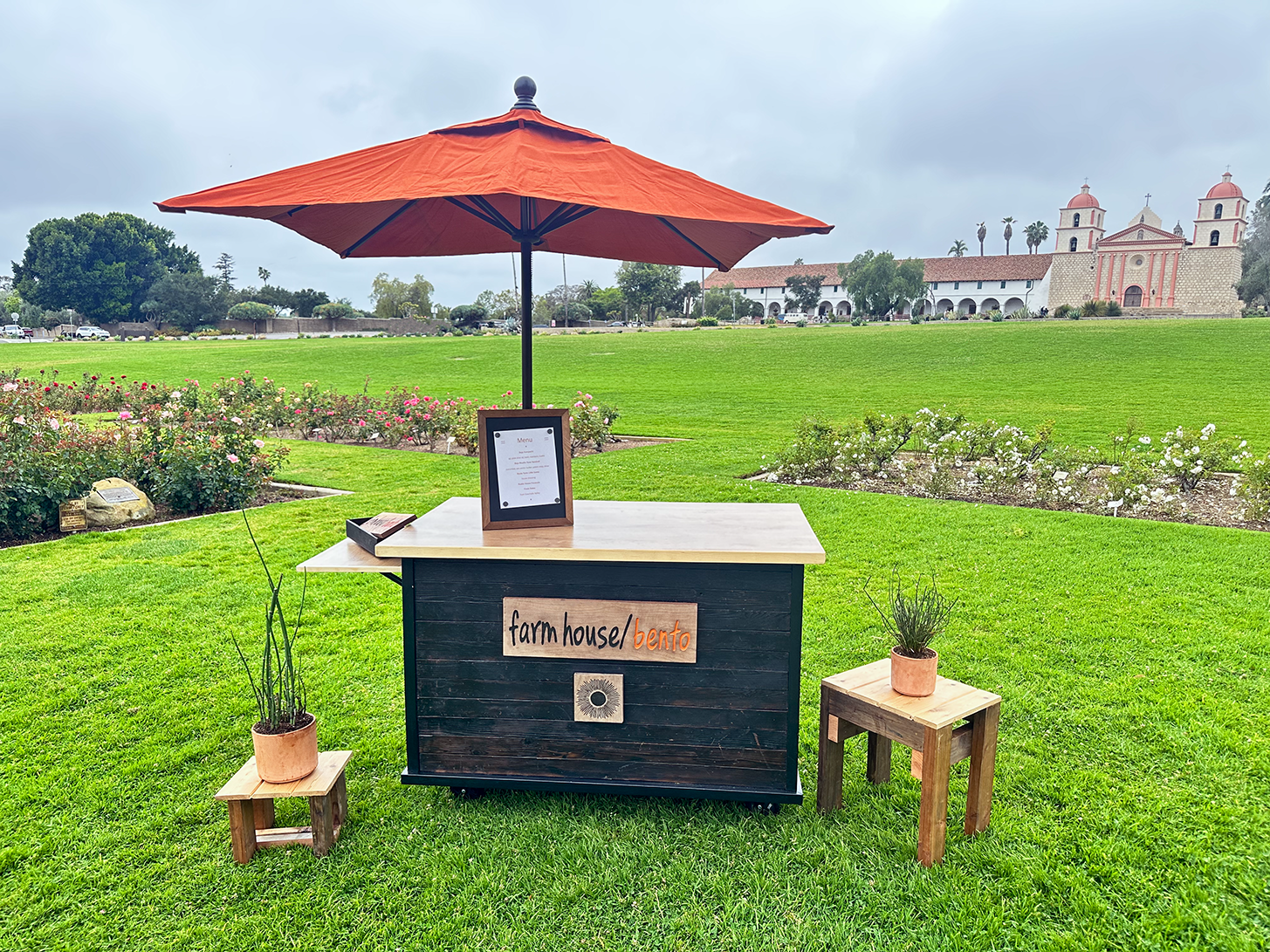 A food stand with an orange umbrella on a green lawn, with a sign saying "farmhouse/bento" on the front, and two small wooden tables with potted plants, one on each side. In the background are flowering bushes, a large building with towers, and a cloudy sky.