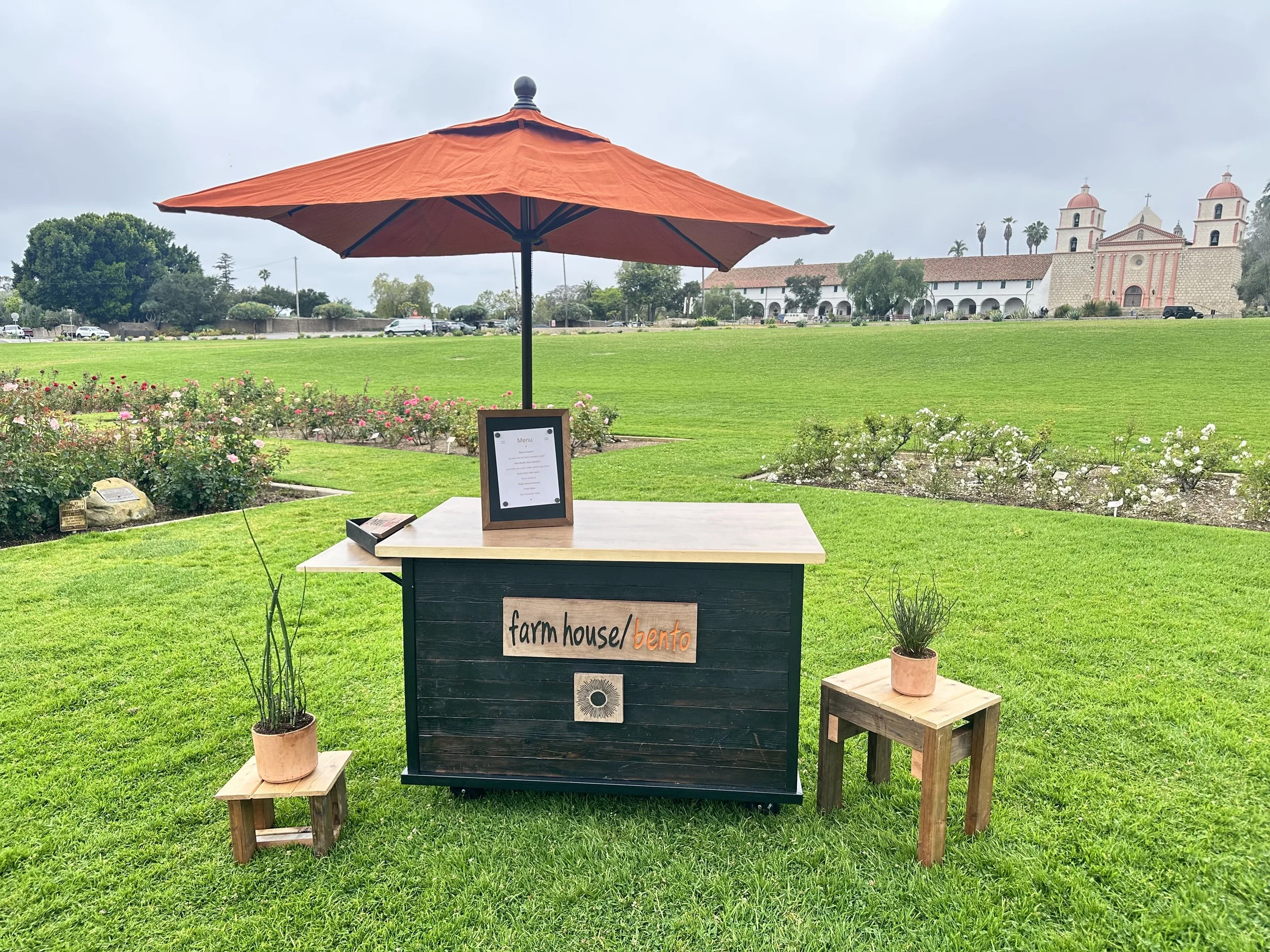 A sign for a farm house and bento, with a black background and white and orange text, placed on a wooden stand in front of a green lawn. There are small potted plants on either side of the sign, and a small table with another potted plant on it. In the background, there is a large grassy field and a historic church with twin bell towers visible under a cloudy sky.