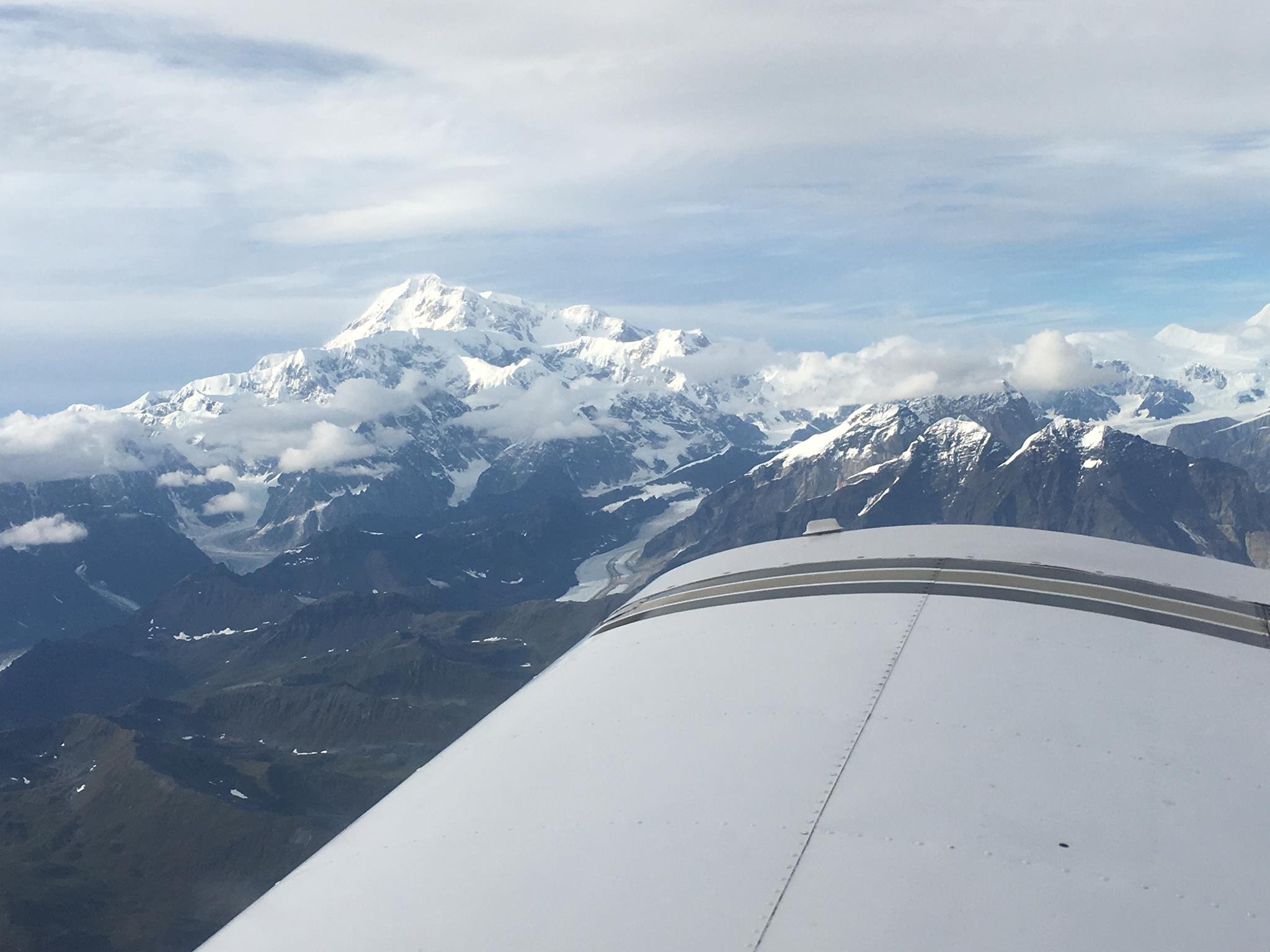 View of snow-covered mountains from an airplane window, with part of the airplane's wing in the foreground.