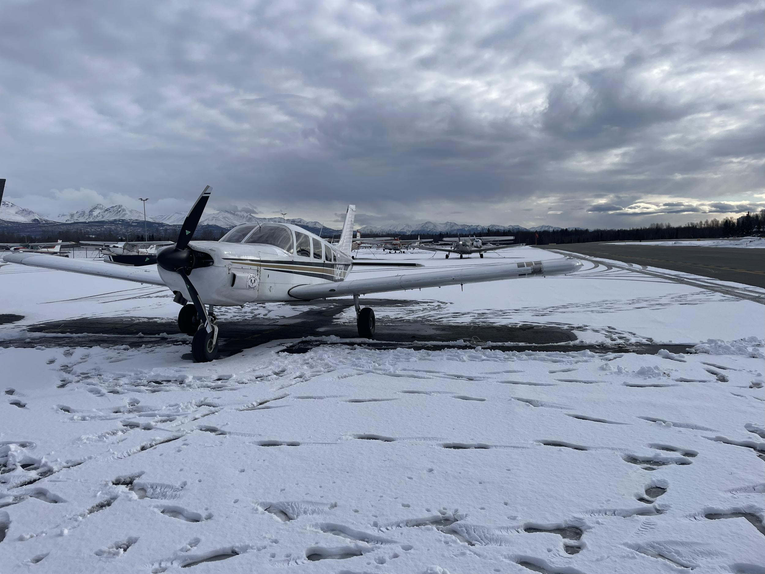 Small white airplane parked on snow-covered tarmac at an airport with mountains in the background and overcast sky.