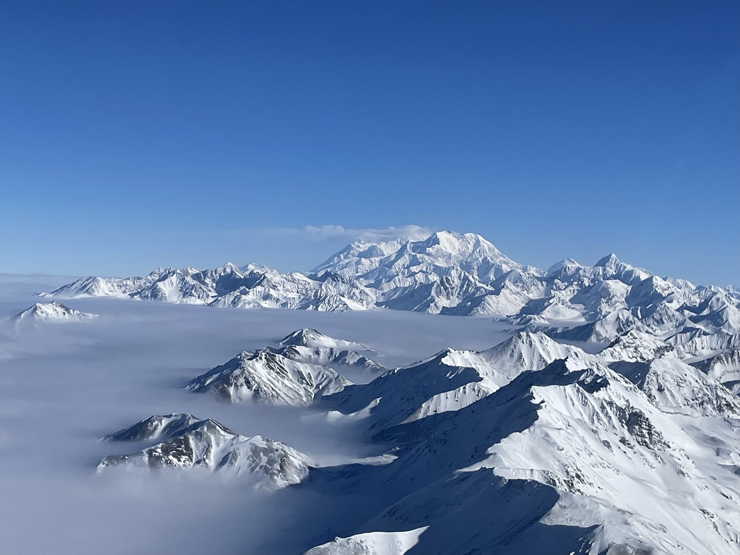 Snow-covered mountain range under a clear blue sky, with some mountains partially shrouded in clouds.