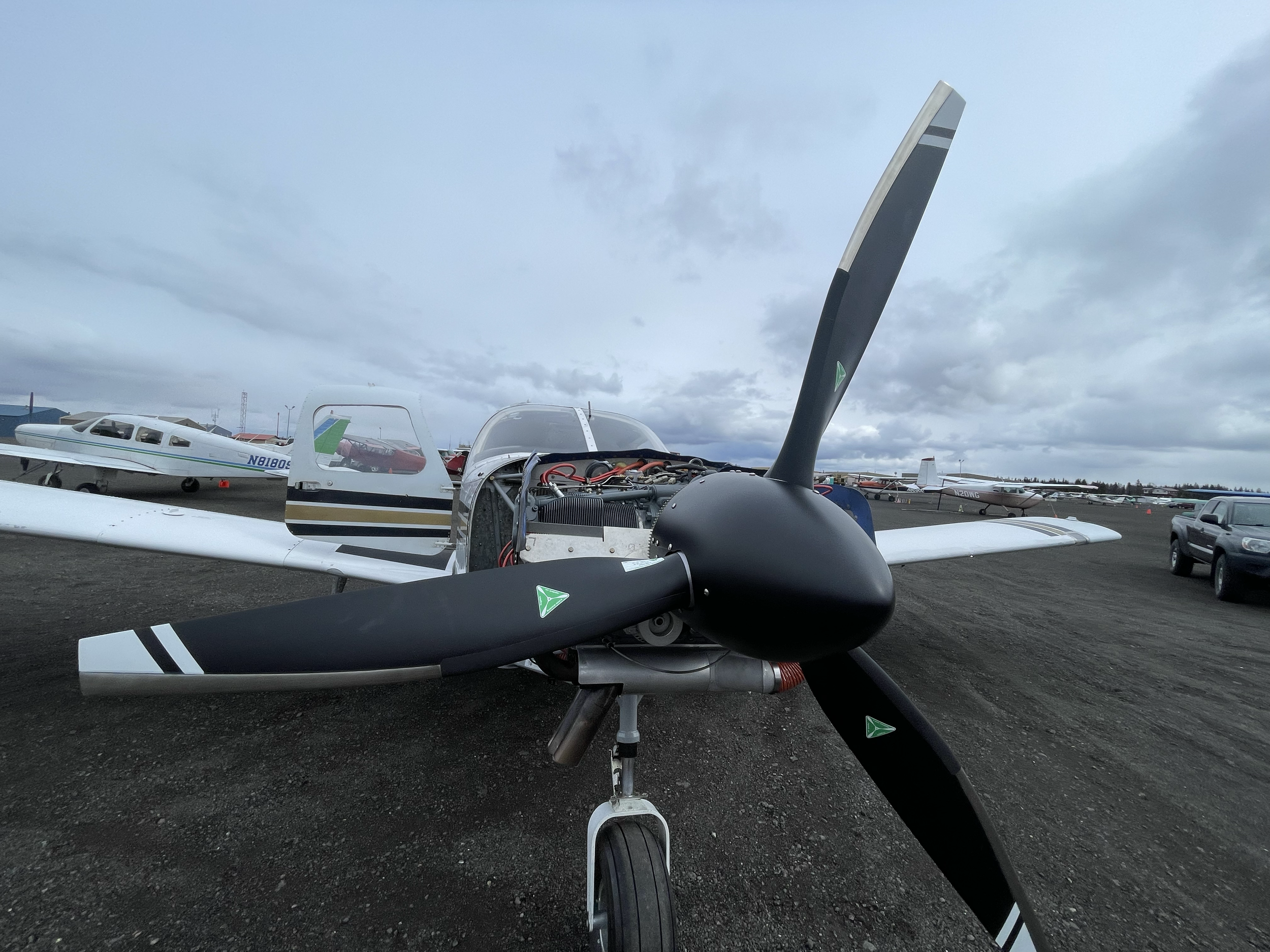 Front view of a small airplane with its engine cover open, parked on a tarmac, with other airplanes and vehicles in the background under cloudy skies.