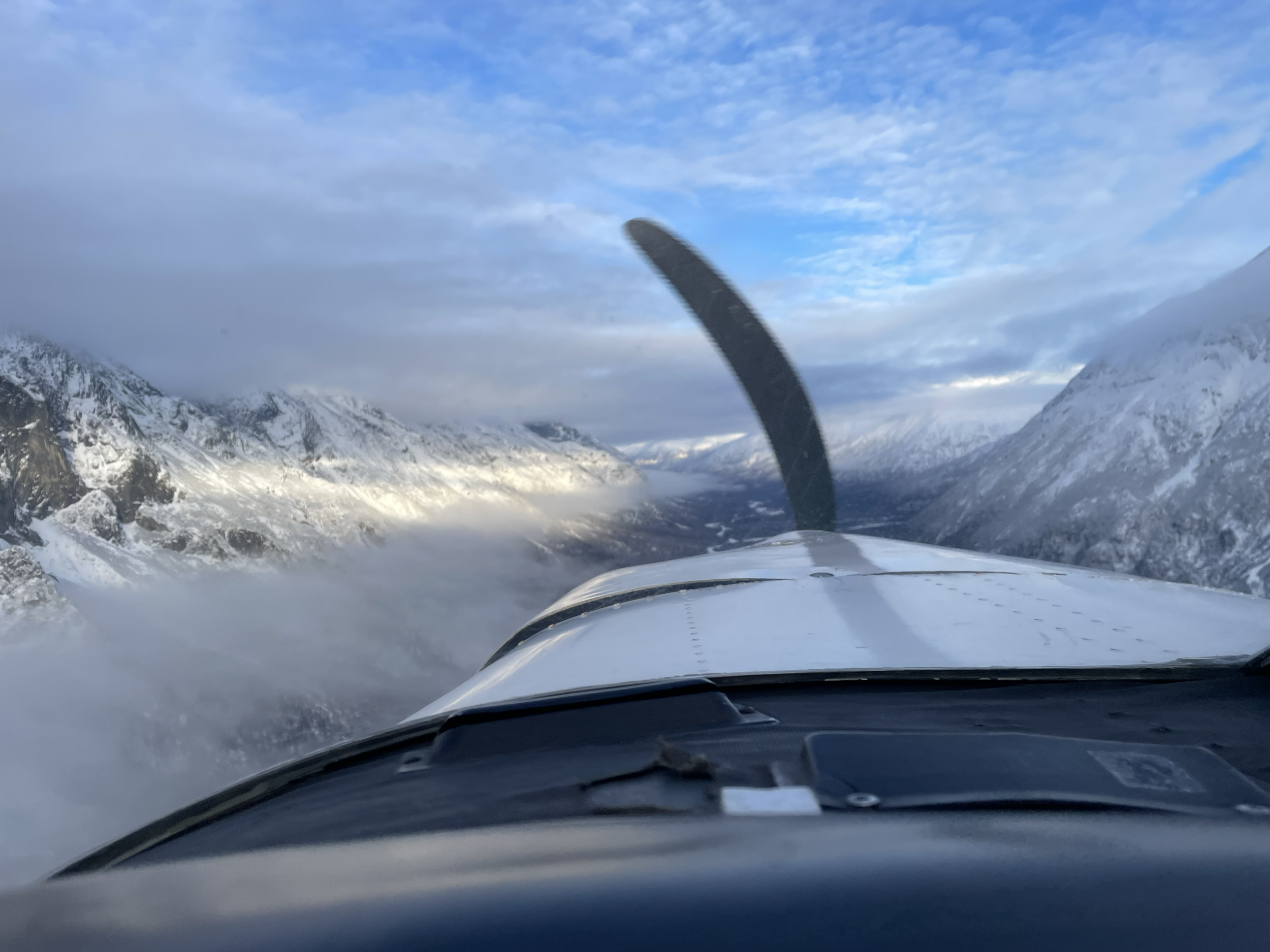 View from inside small airplane cockpit flying over snow-covered mountains with cloudy sky.