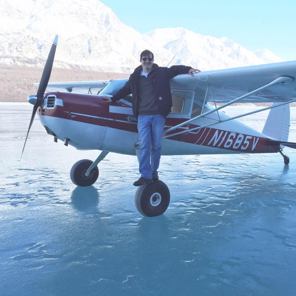 Person standing on the wheel of a small airplane on an icy surface with mountains in the background.