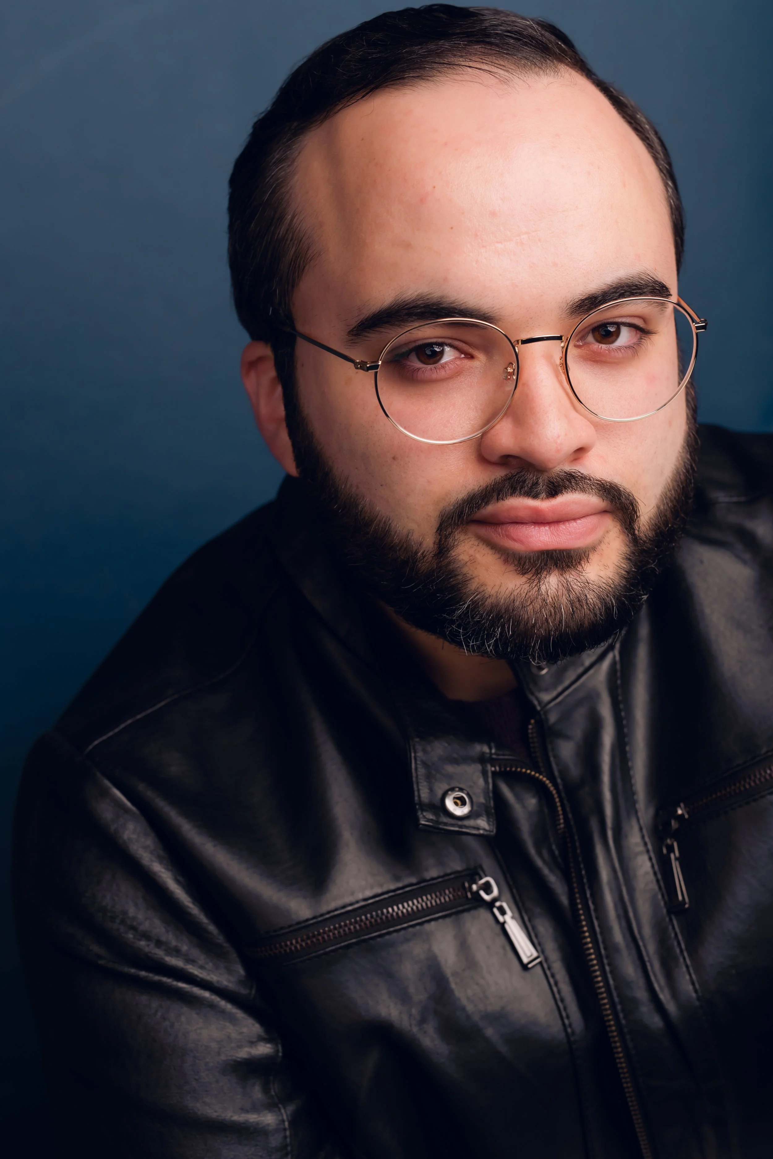 Close-up portrait of a young man with glasses, a beard, and dark hair, wearing a black leather jacket, against a blue background.
