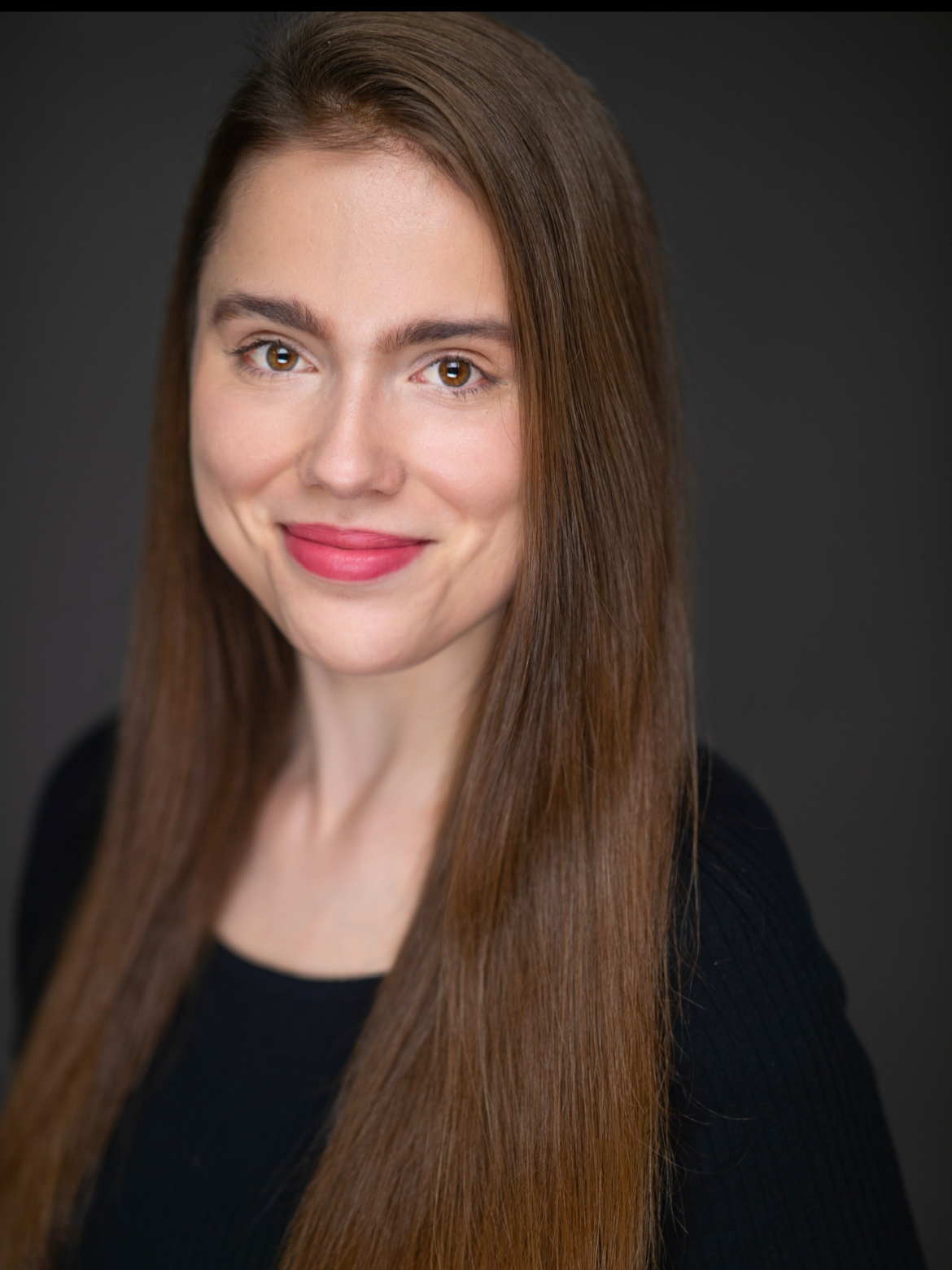 A woman with long brown hair, light skin, and wearing a black top, smiling at the camera against a dark background.