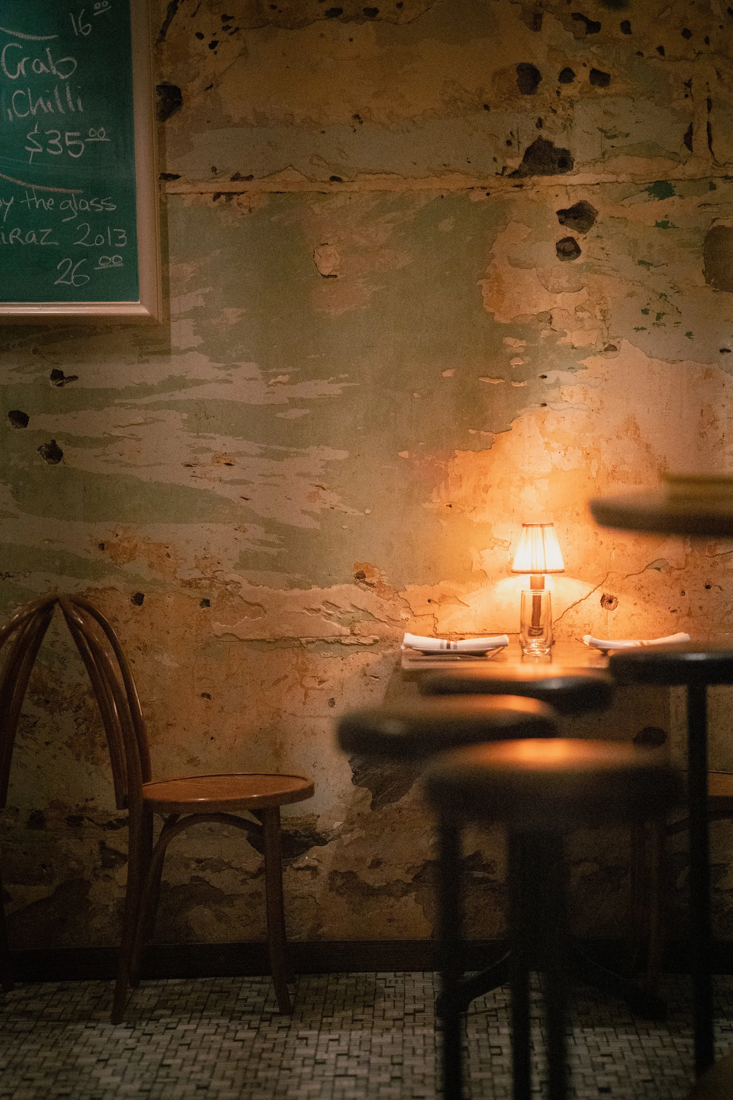 Rustic restaurant interior with a weathered, peeling wall, a small table with a lamp providing warm light, and empty chairs.
