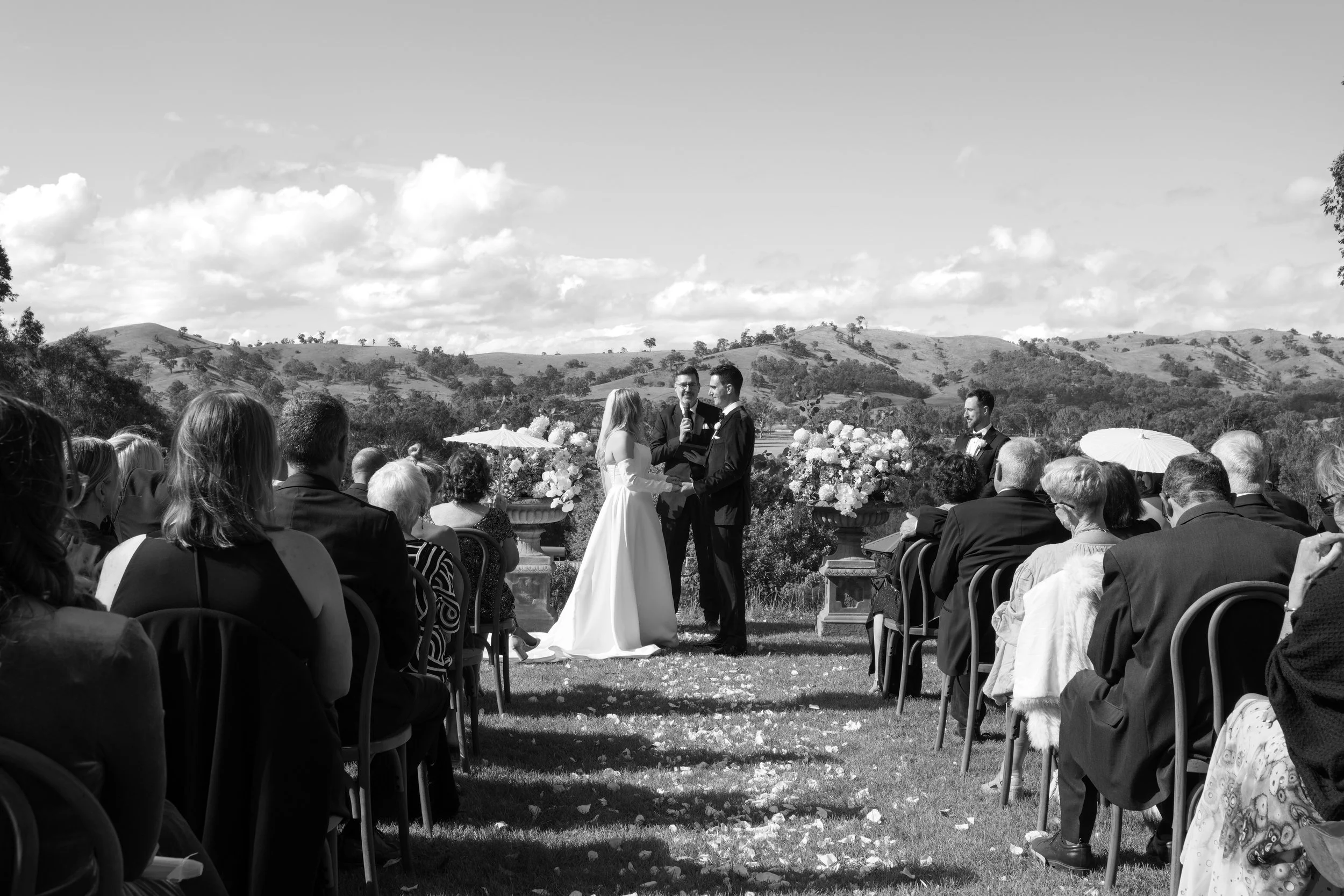 A black and white photograph of an outdoor wedding ceremony with a bride and groom holding hands and exchanging vows, surrounded by seated guests, with a scenic landscape of hills and sky in the background.