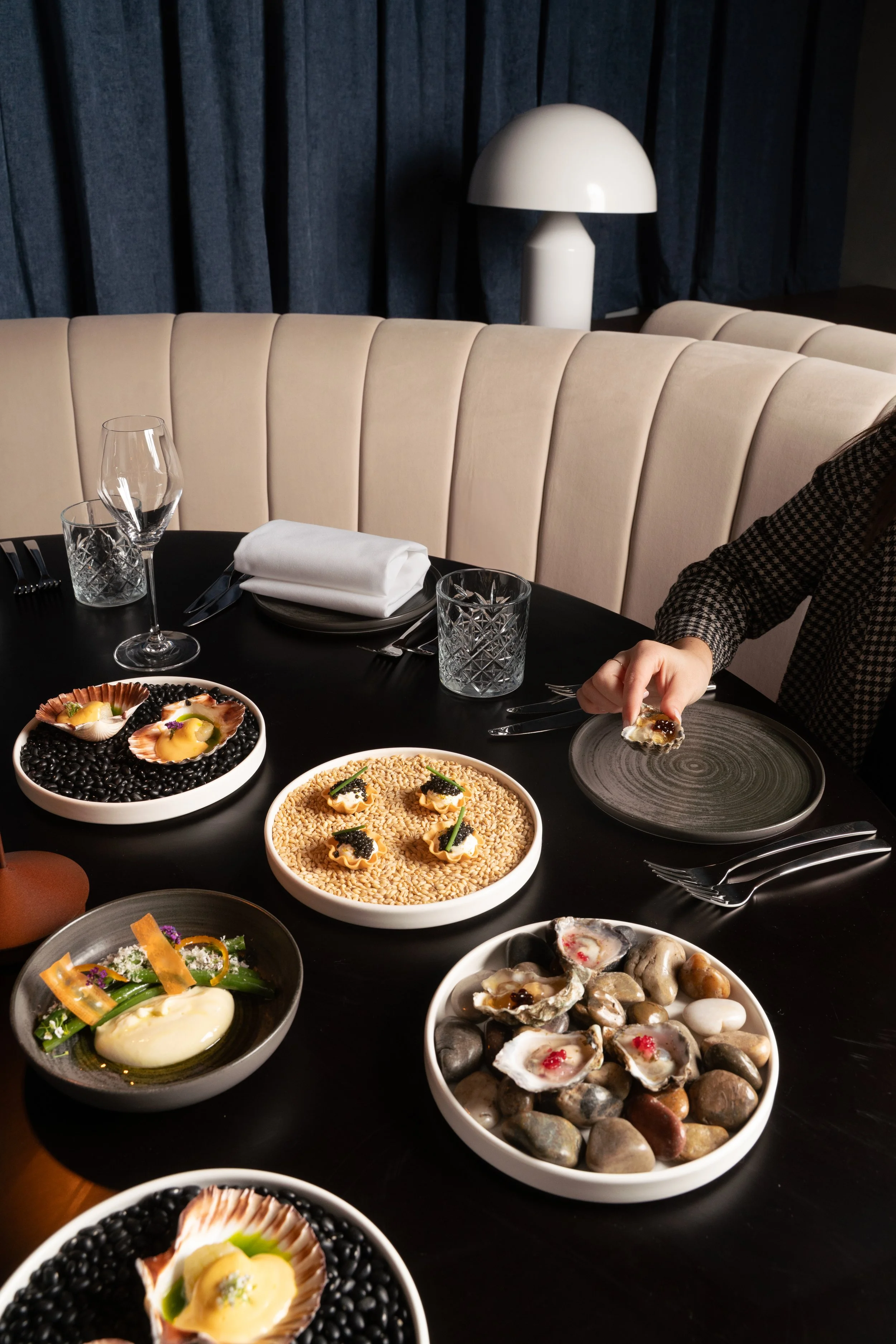 Table set with various appetizers and seafood dishes, including oysters, caviar, and appetizers garnished with herbs, on a black table with drinking glasses and silverware.