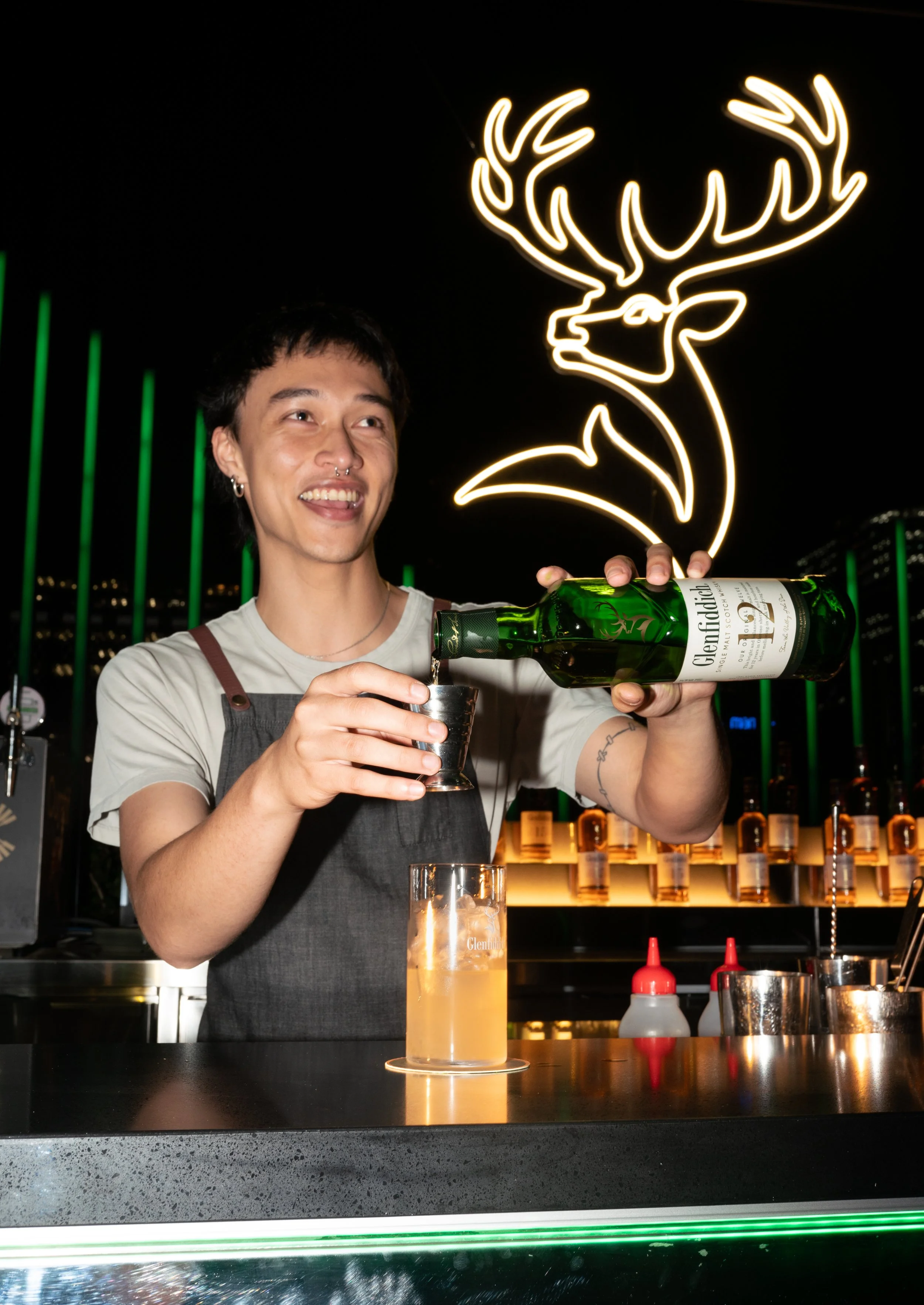A bartender pours a drink from a green bottle of Glenfiddich whiskey at a bar, with a neon reindeer sign in the background.
