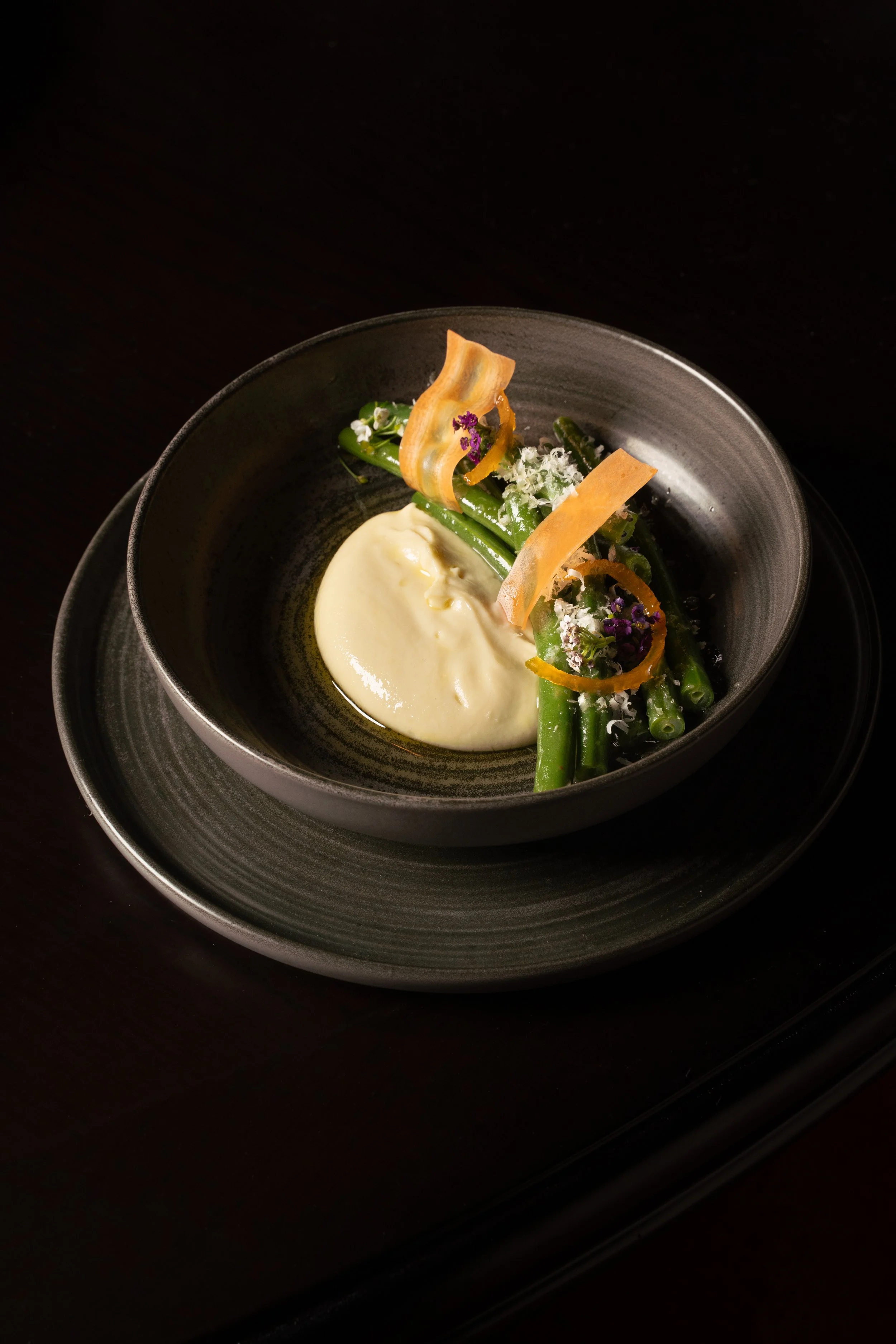 A plated gourmet dish with green beans, a dollop of creamy sauce, garnished with crispy vegetable chips, edible purple flowers, and grated cheese, served in a black bowl on a matching black plate.
