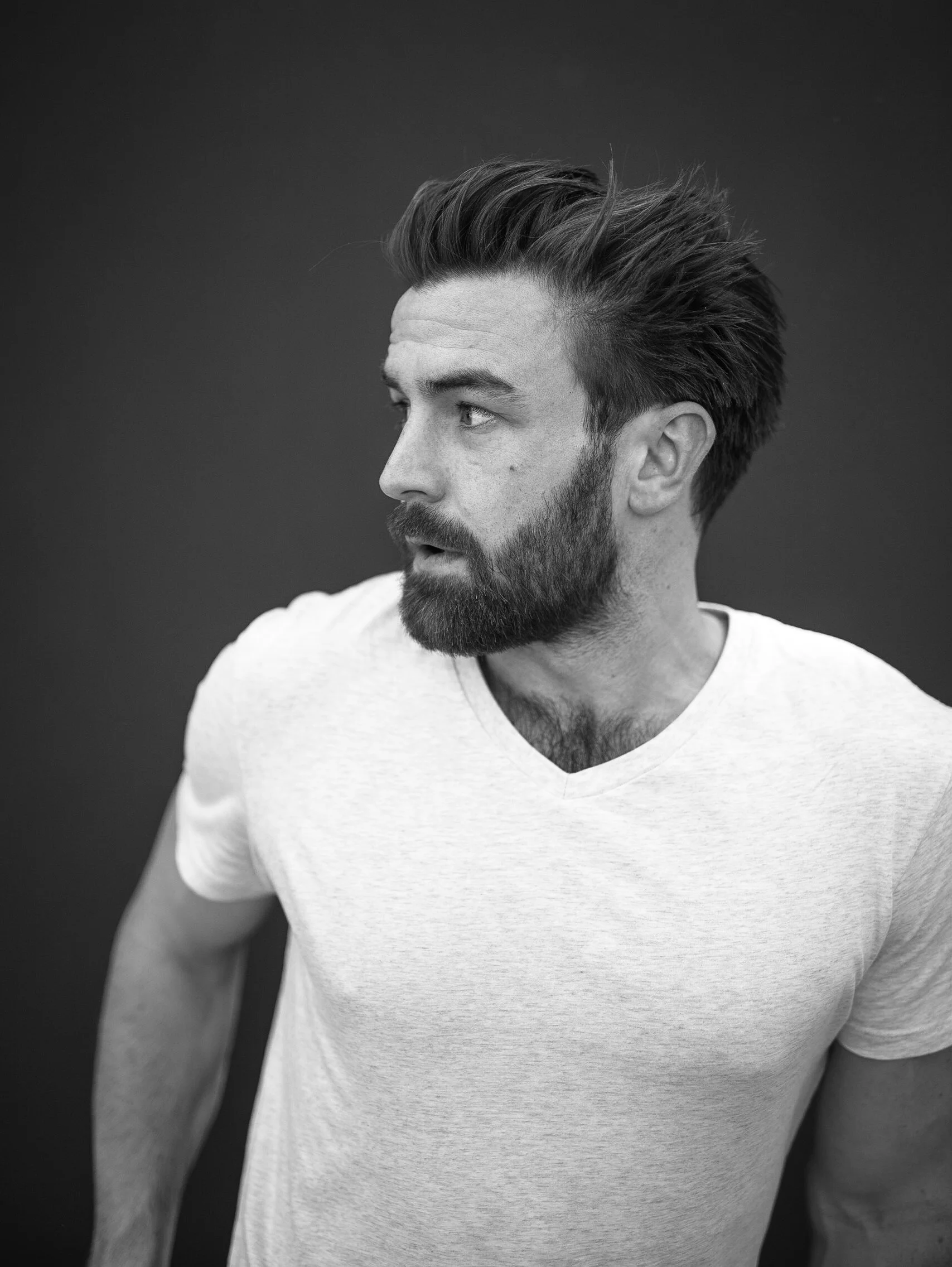 Black and white portrait of a man with a beard and styled hair, wearing a light-colored t-shirt, looking to his left with a neutral expression.