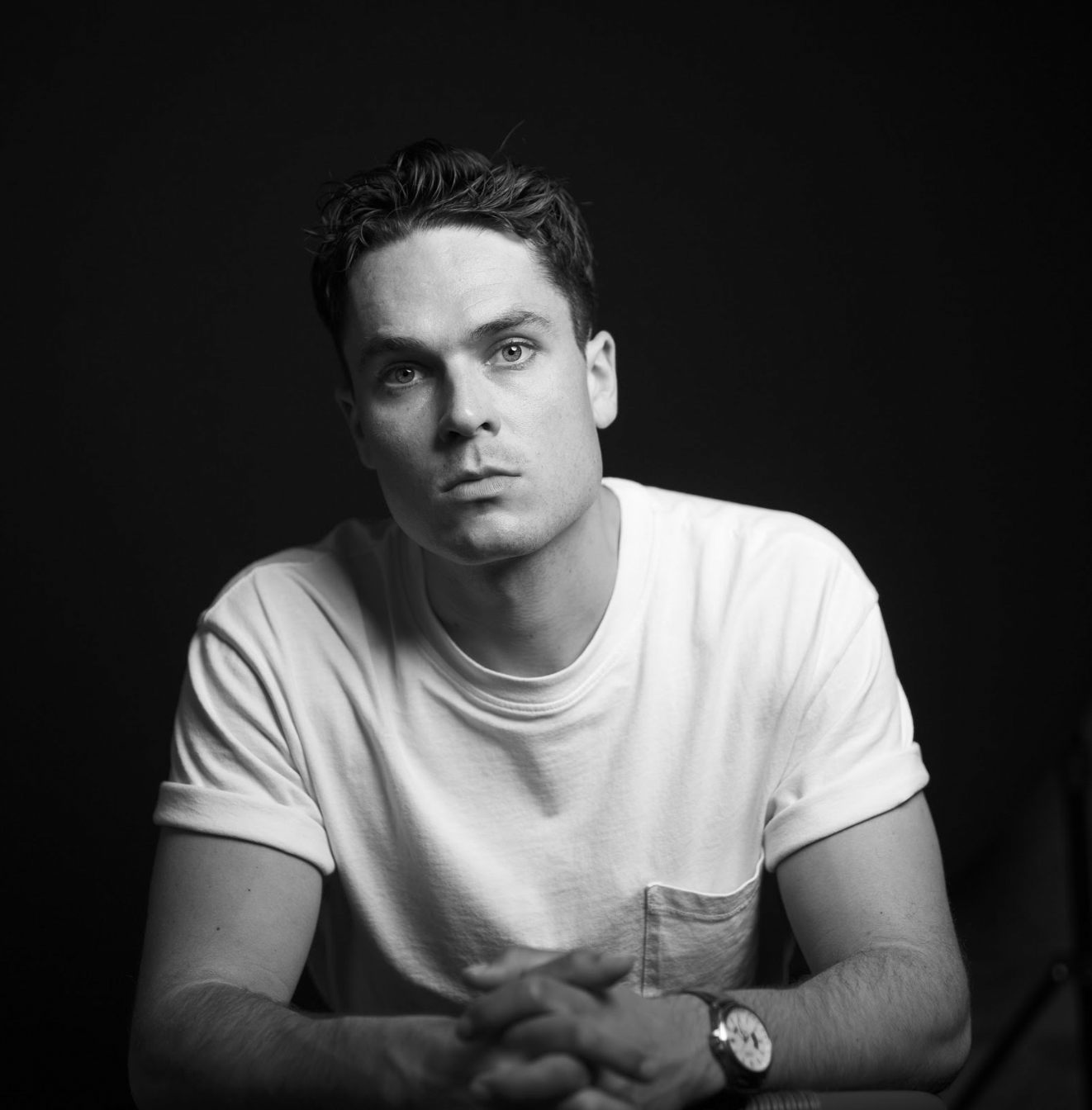 Black and white portrait of a young man with short, dark hair, wearing a white t-shirt and a watch, sitting against a dark background with hands clasped.