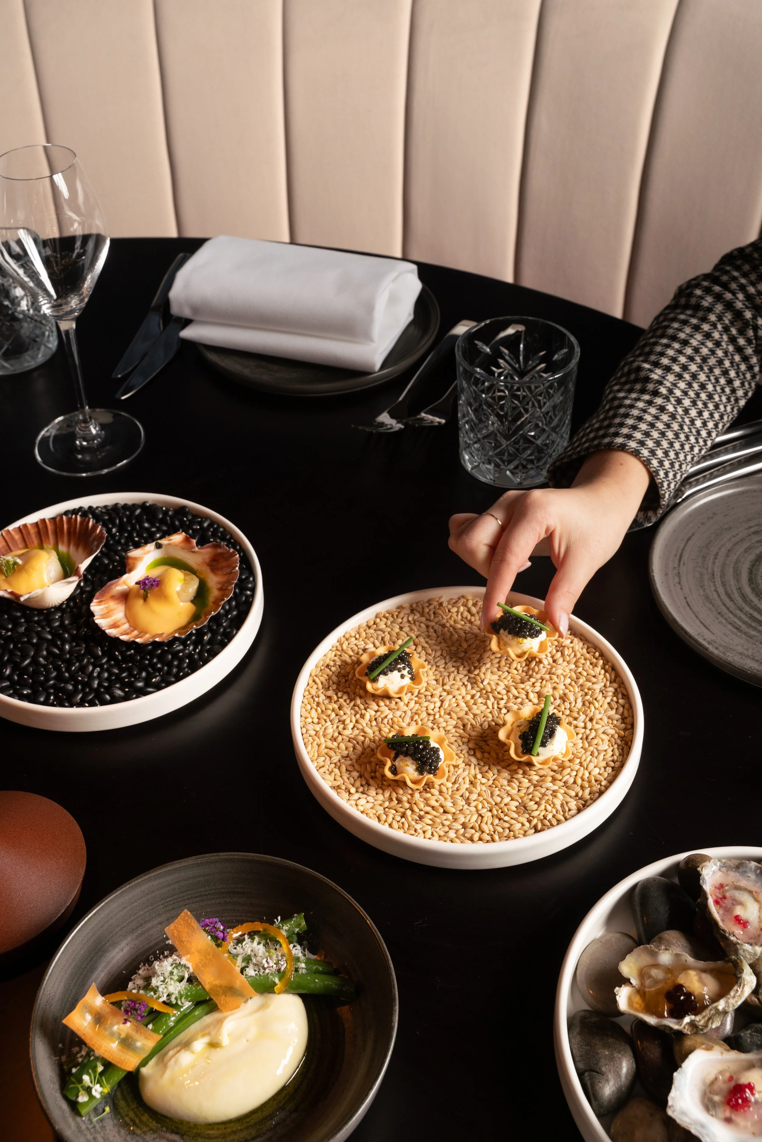 Restaurant table with assorted gourmet dishes, including a large bowl of small pastries topped with caviar and garnishes, a dish with green vegetables, and another with oysters, set with glassware, silverware, and a folded napkin.