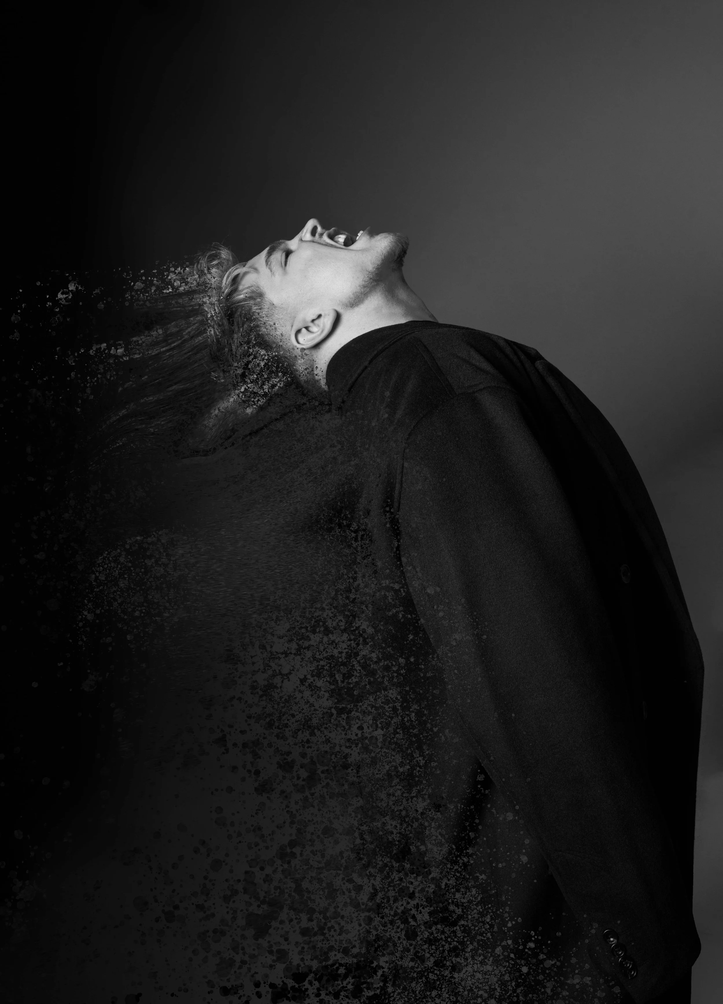 A black and white photo of a man with his head tilted back, mouth open, and hair flying back as if he is shaking his head. The background is dark, and there is a dynamic, splattered effect around his head.