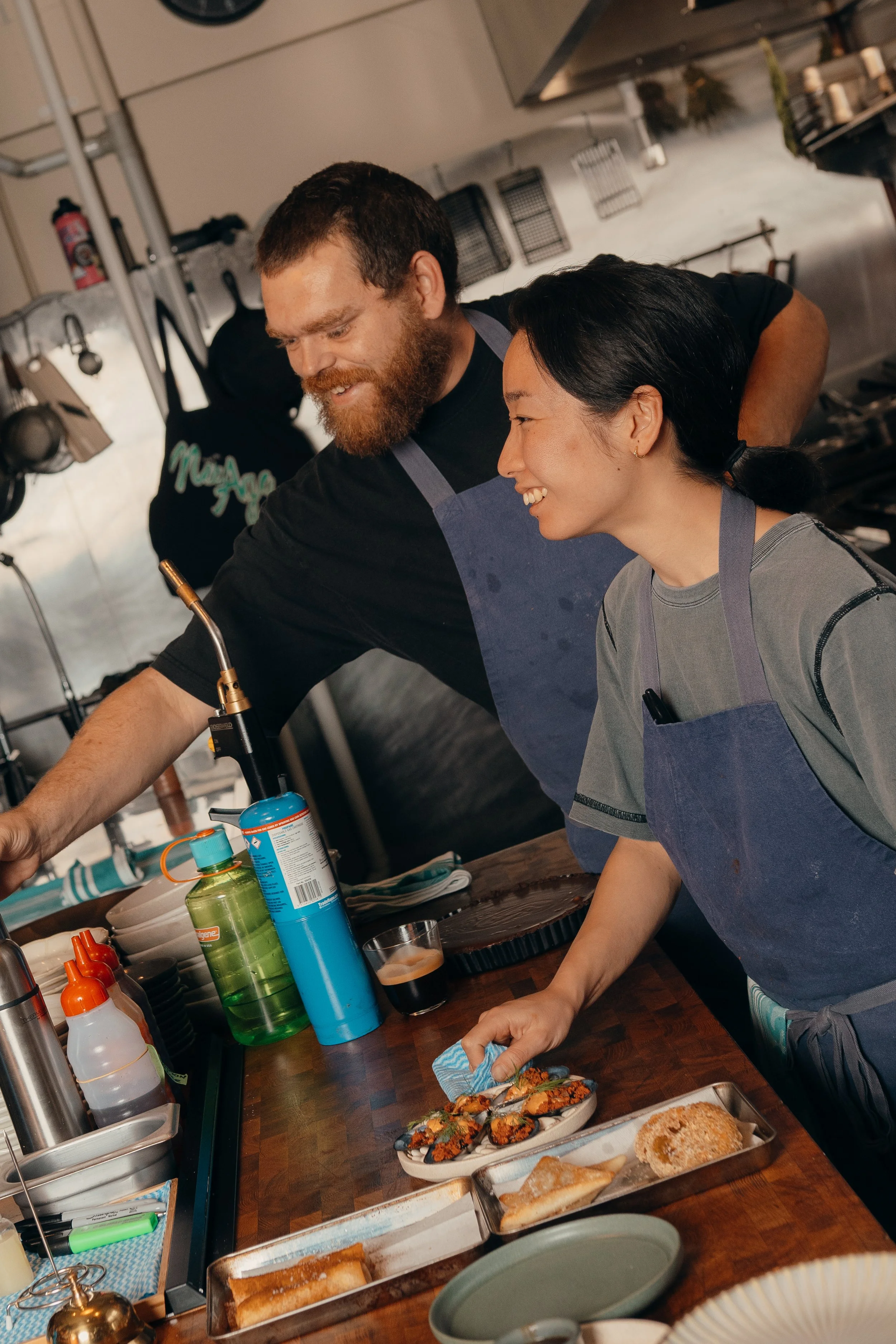 Two chefs, a man with a beard and a woman with black hair, working together in a restaurant kitchen, preparing and plating dishes on a wooden counter.
