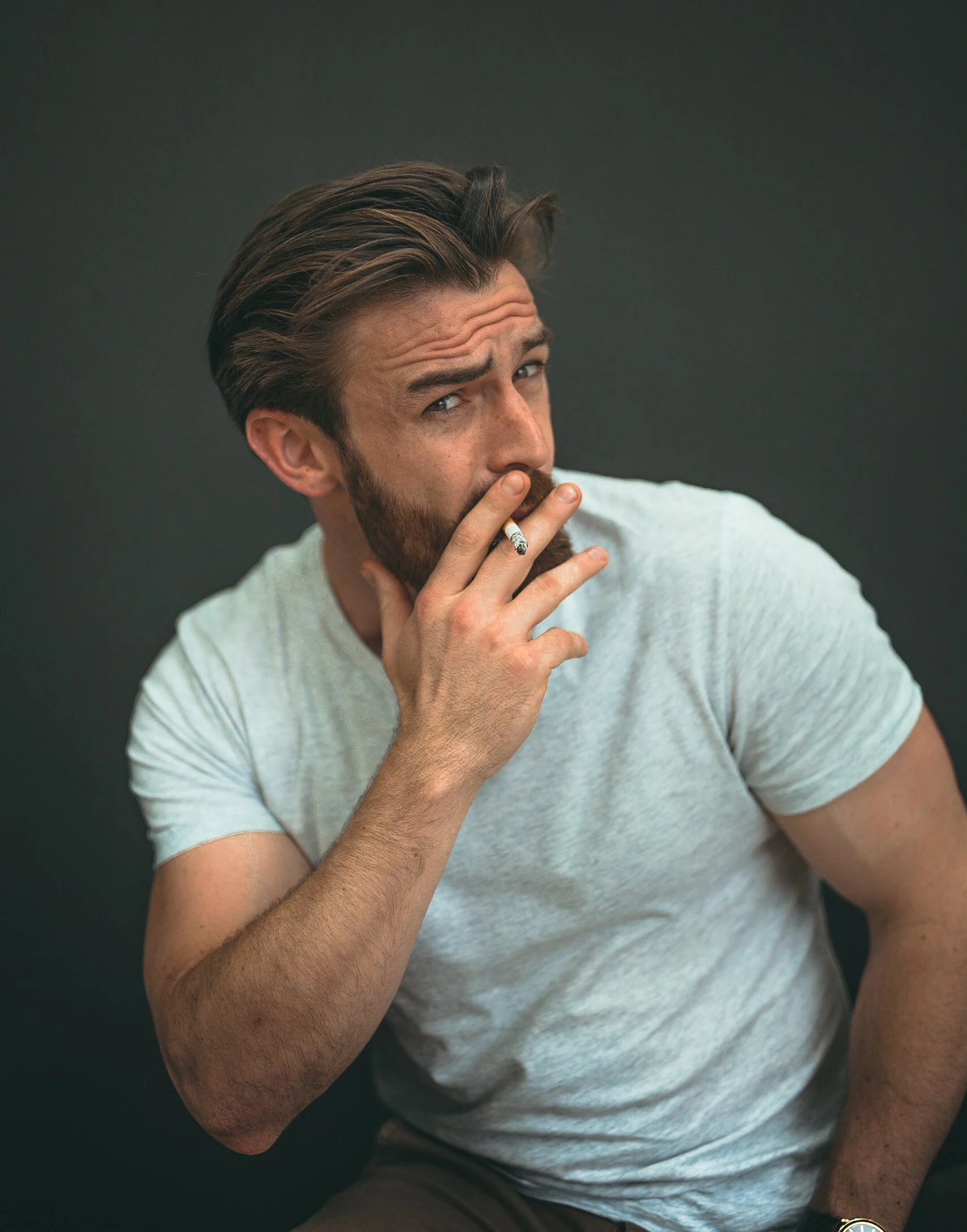 A man with brown hair and a beard, wearing a light gray t-shirt, smokes a cigarette against a dark background.