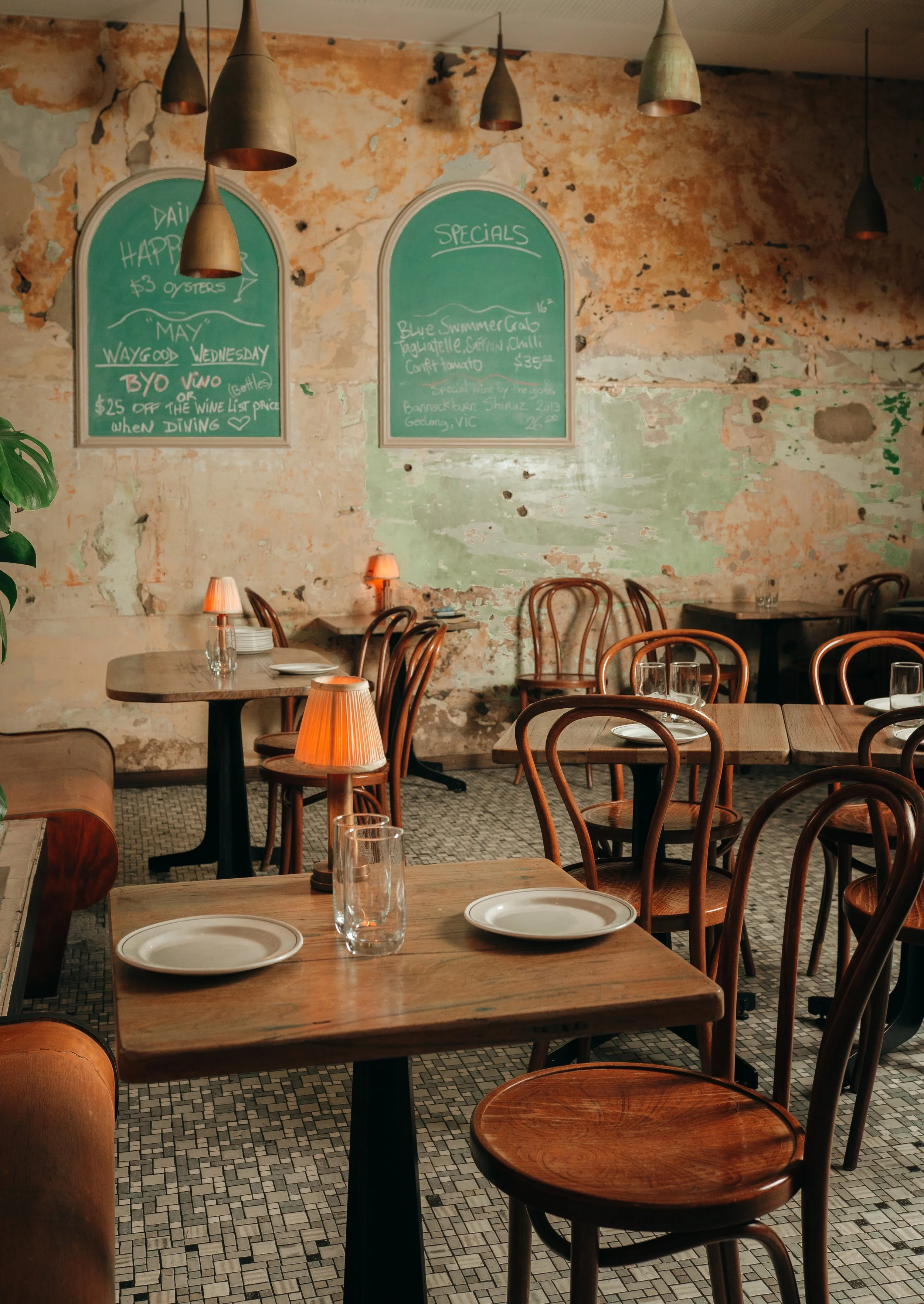 Empty restaurant with wooden tables and chairs, chalkboards with specials on a distressed wall, and pendant lighting.