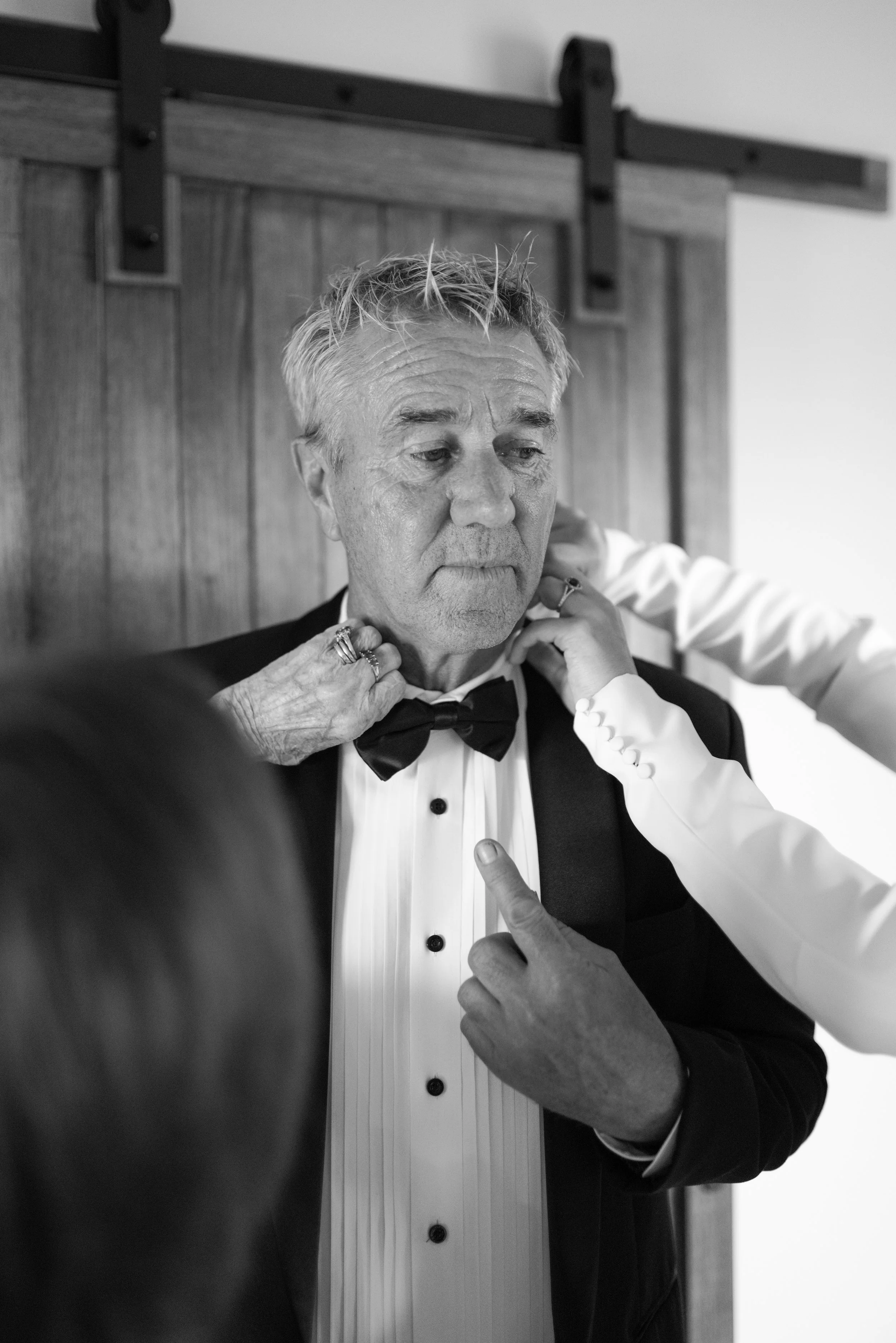 A man in a tuxedo with a bowtie getting dressed, assisting him with his tuxedo. Black and white photo.