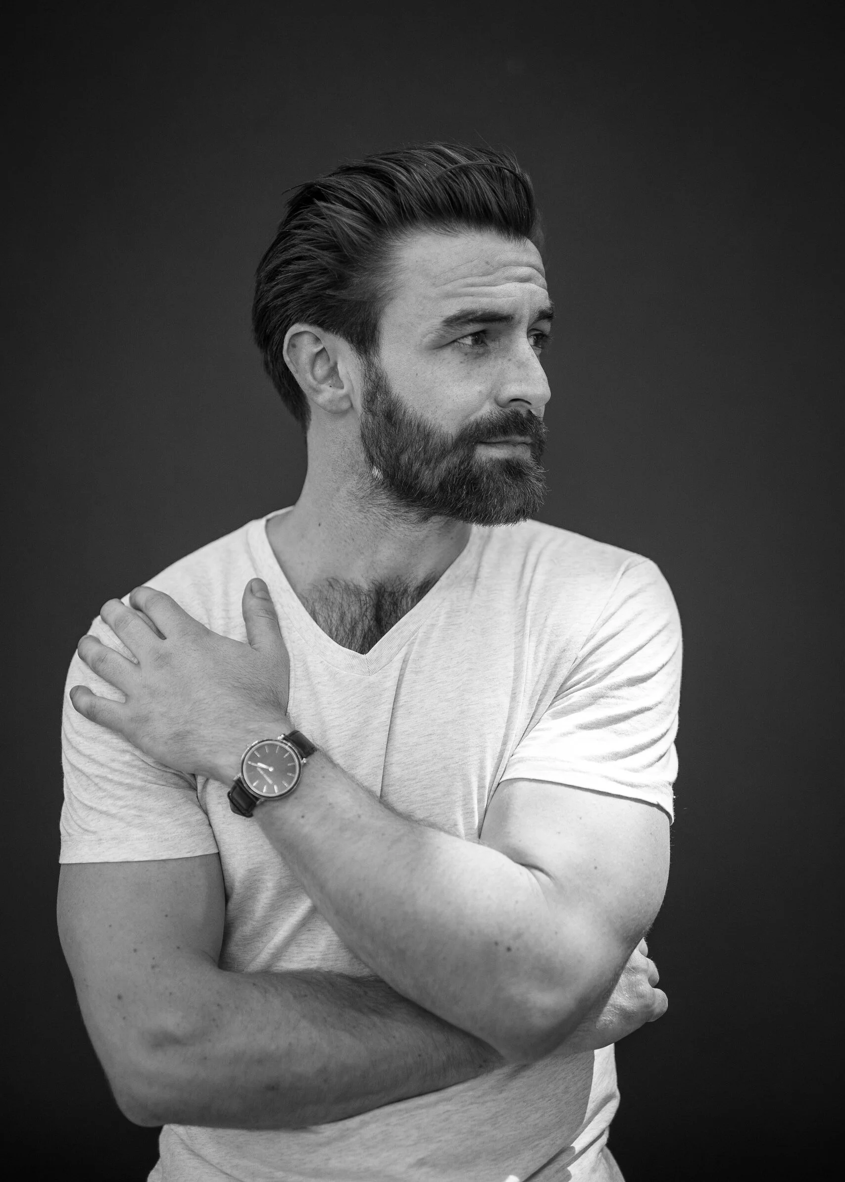 Black and white portrait of a man with a beard and styled hair, wearing a watch and a light-colored t-shirt, looking to the side with arms crossed against a dark background.