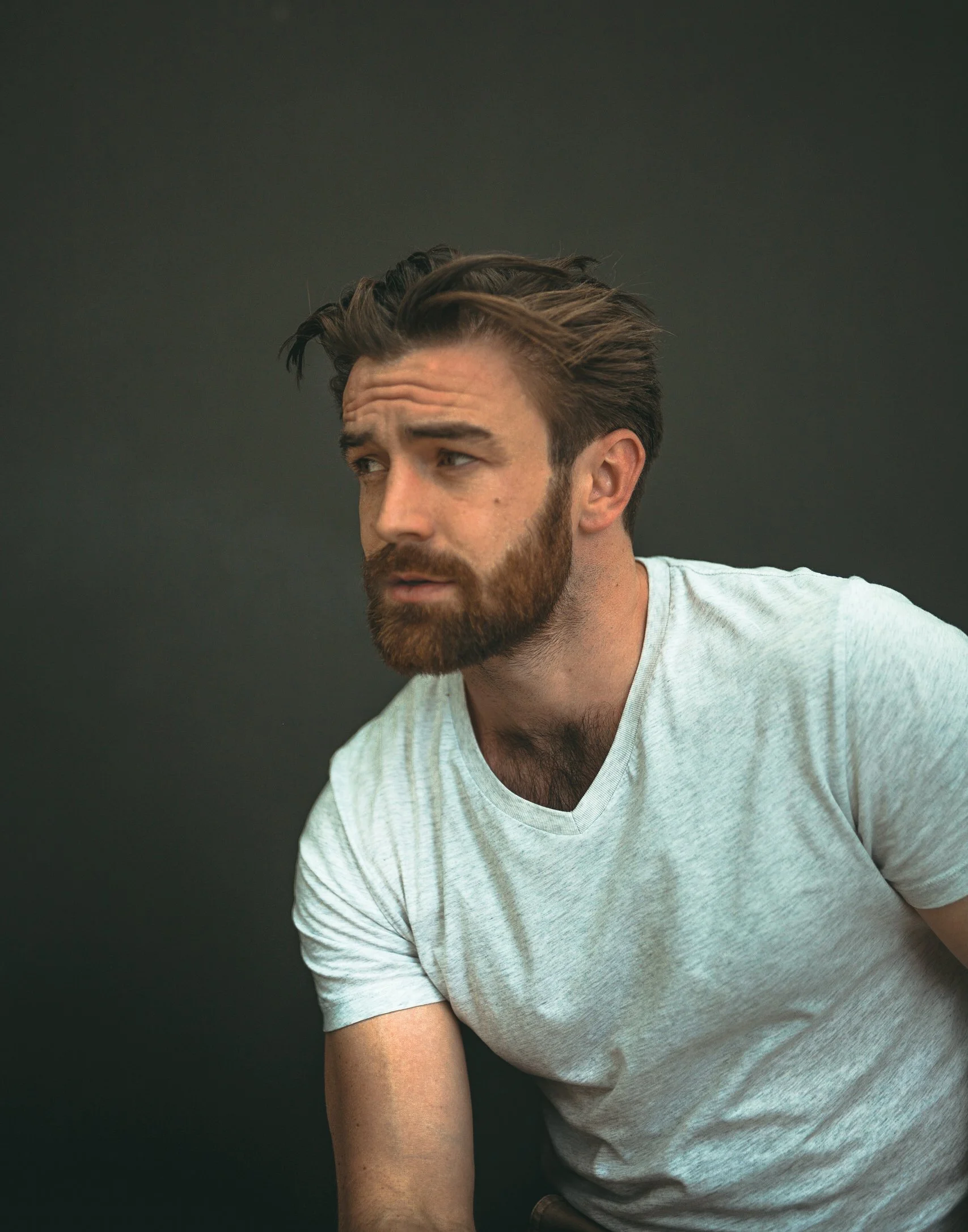 A man with a beard and styled hair looking to the side against a dark background, wearing a light gray T-shirt.