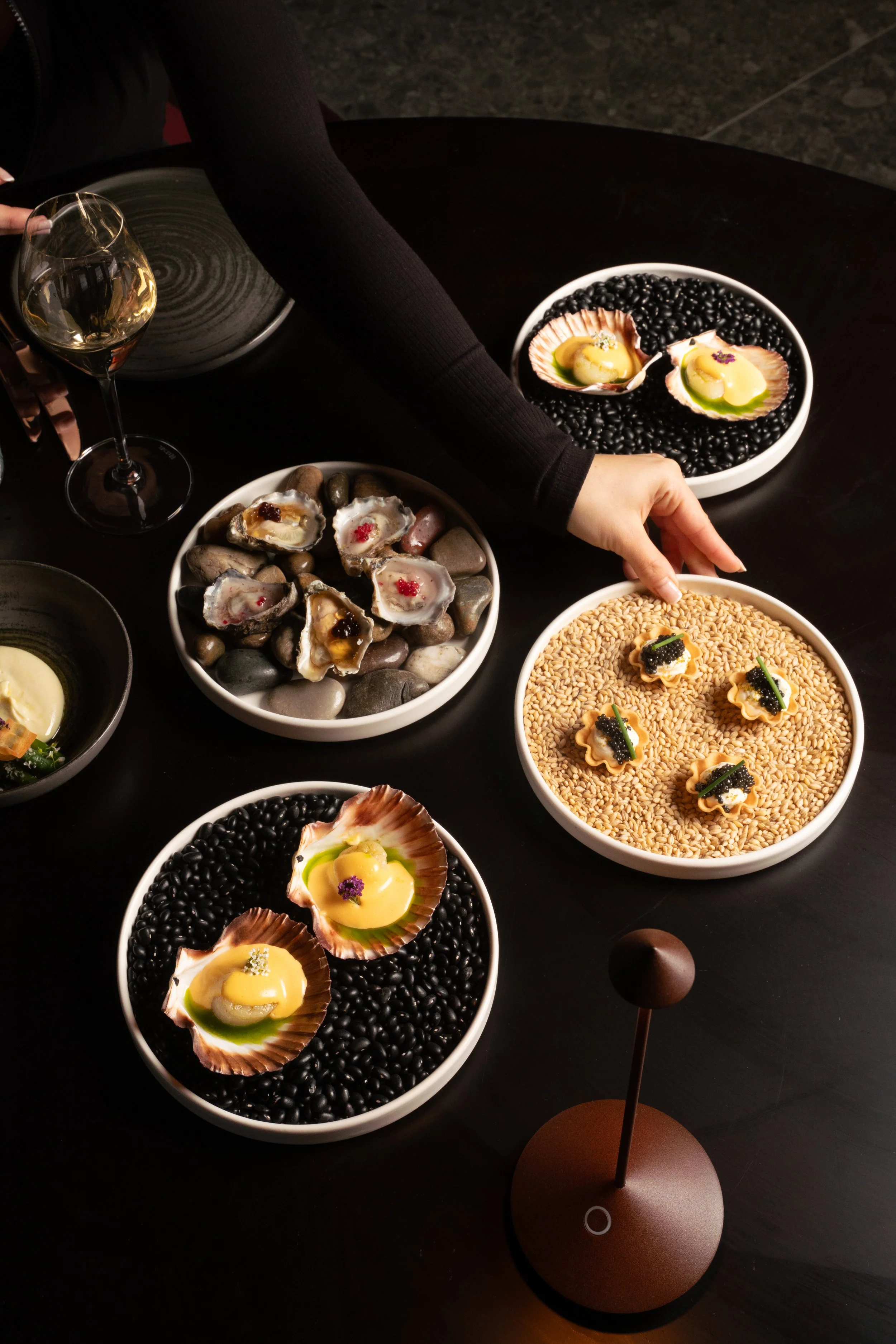 Assorted gourmet dishes on a black table, including shells with yellow filling topped with herbs, bowls of black beans, gray rocks with seafood, and a glass of white wine.
