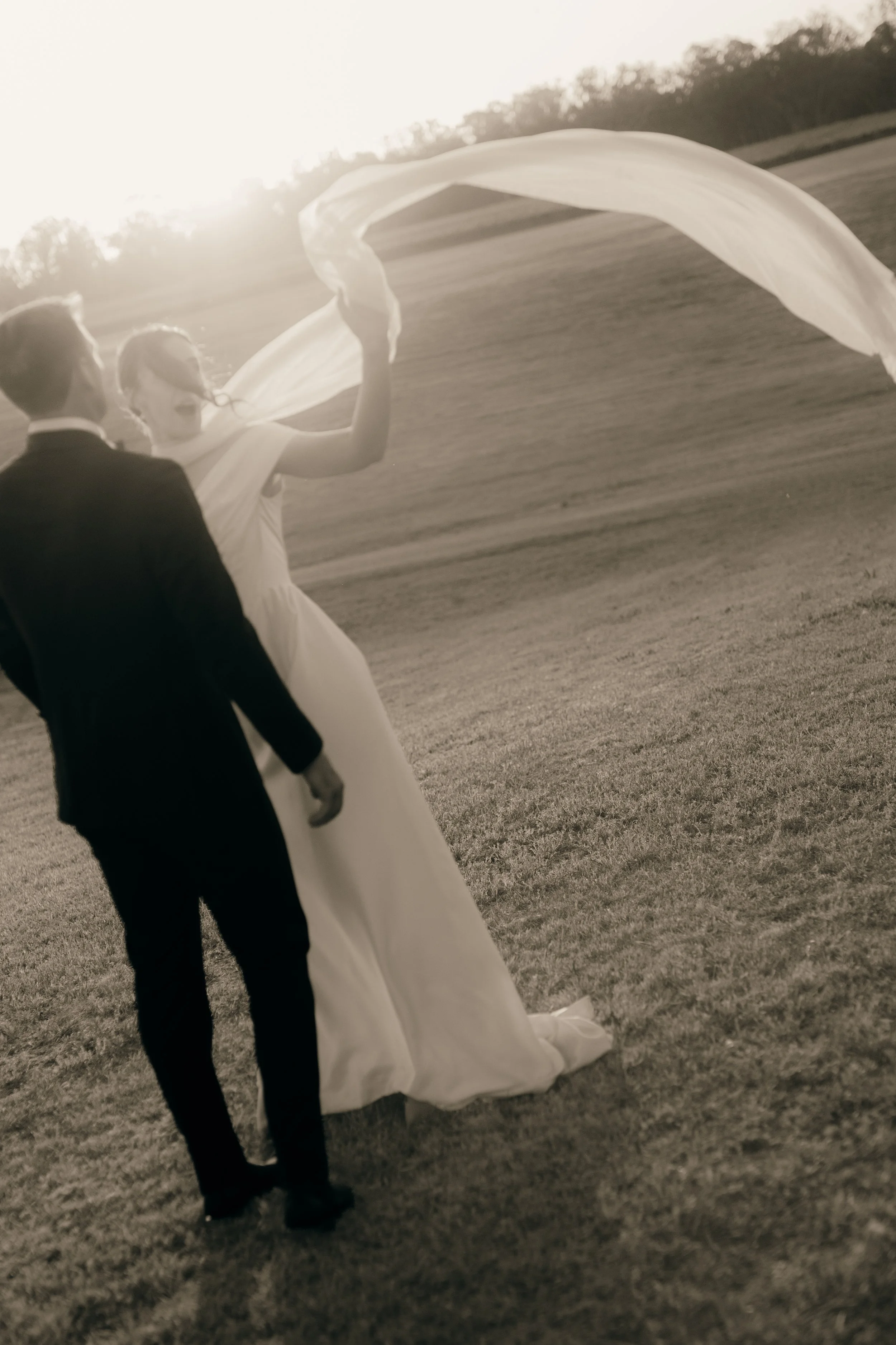 A couple dressed in wedding attire, with the bride holding a flowing cloth, standing on a grassy field during sunset.