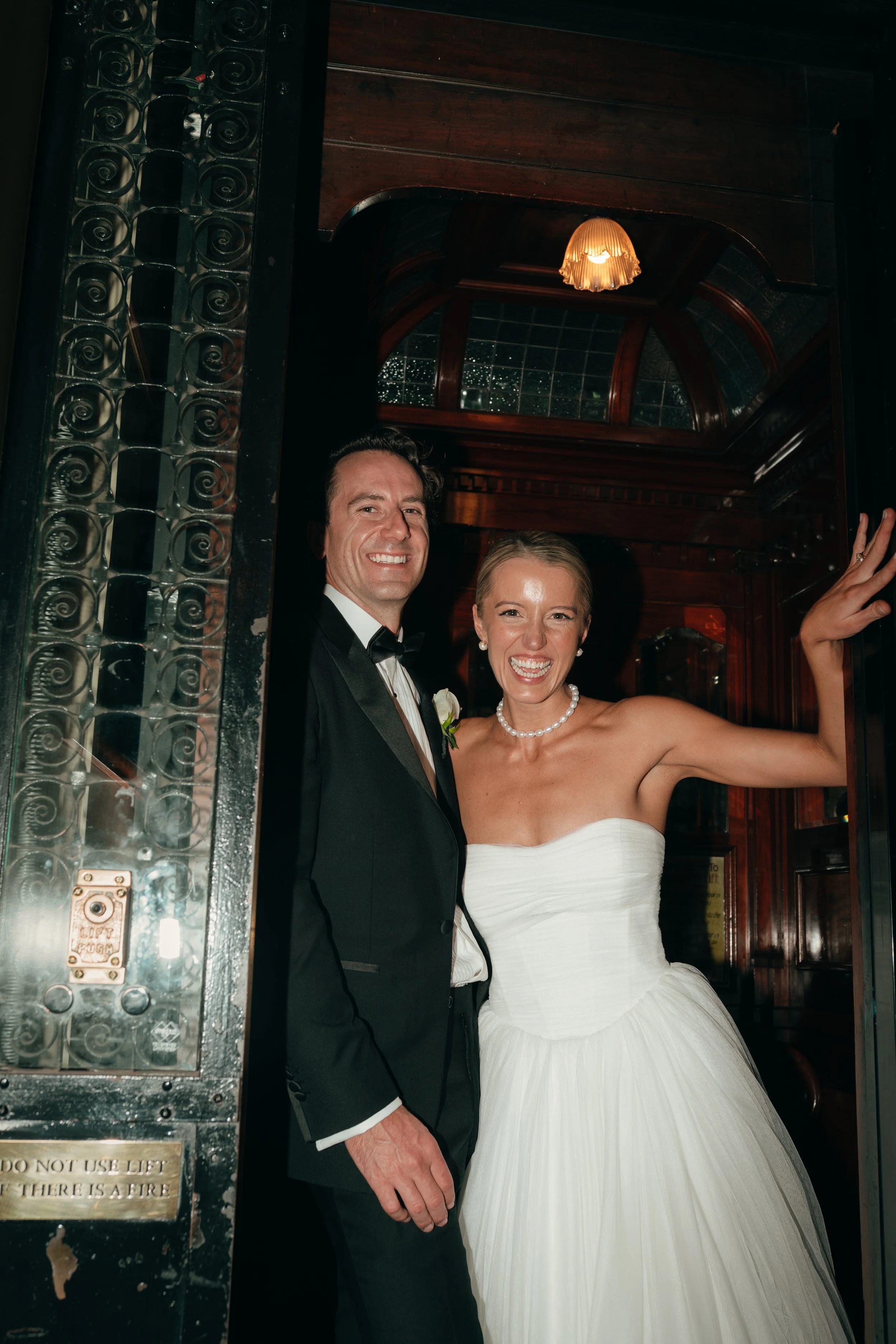 Happy bride and groom smiling and waving, dressed in wedding attire, standing in the doorway of a vintage elevator with dark wood paneling and decorative glass and metal details.