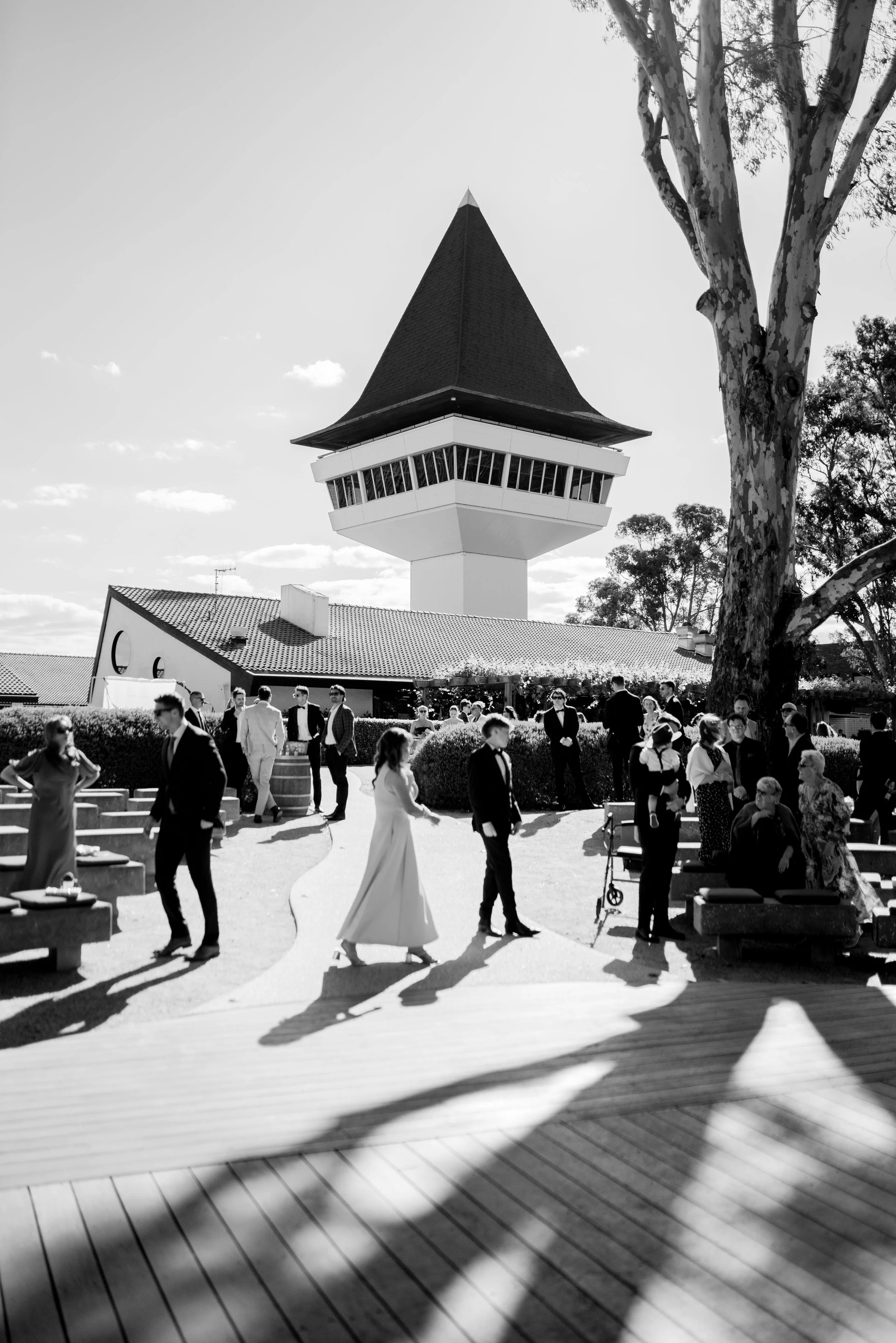 People dressed in formal attire gathered outdoors near a unique tower with a conical roof, trees, and a clear sky.