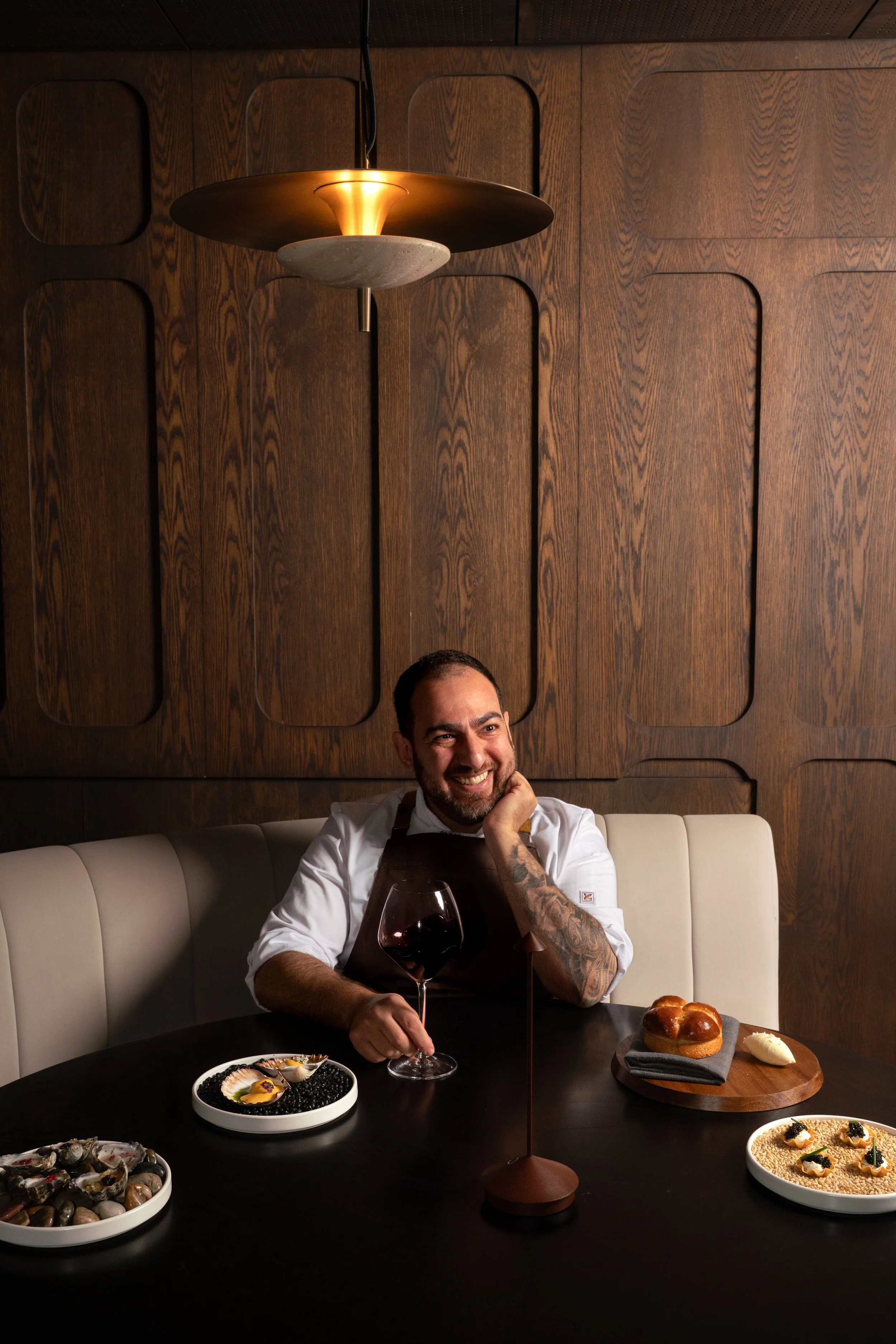A smiling chef with a beard and tattooed arm sitting at a dark table with a glass of red wine, plates of assorted appetizers, and bread. The background features a wooden wall and modern pendant lighting.