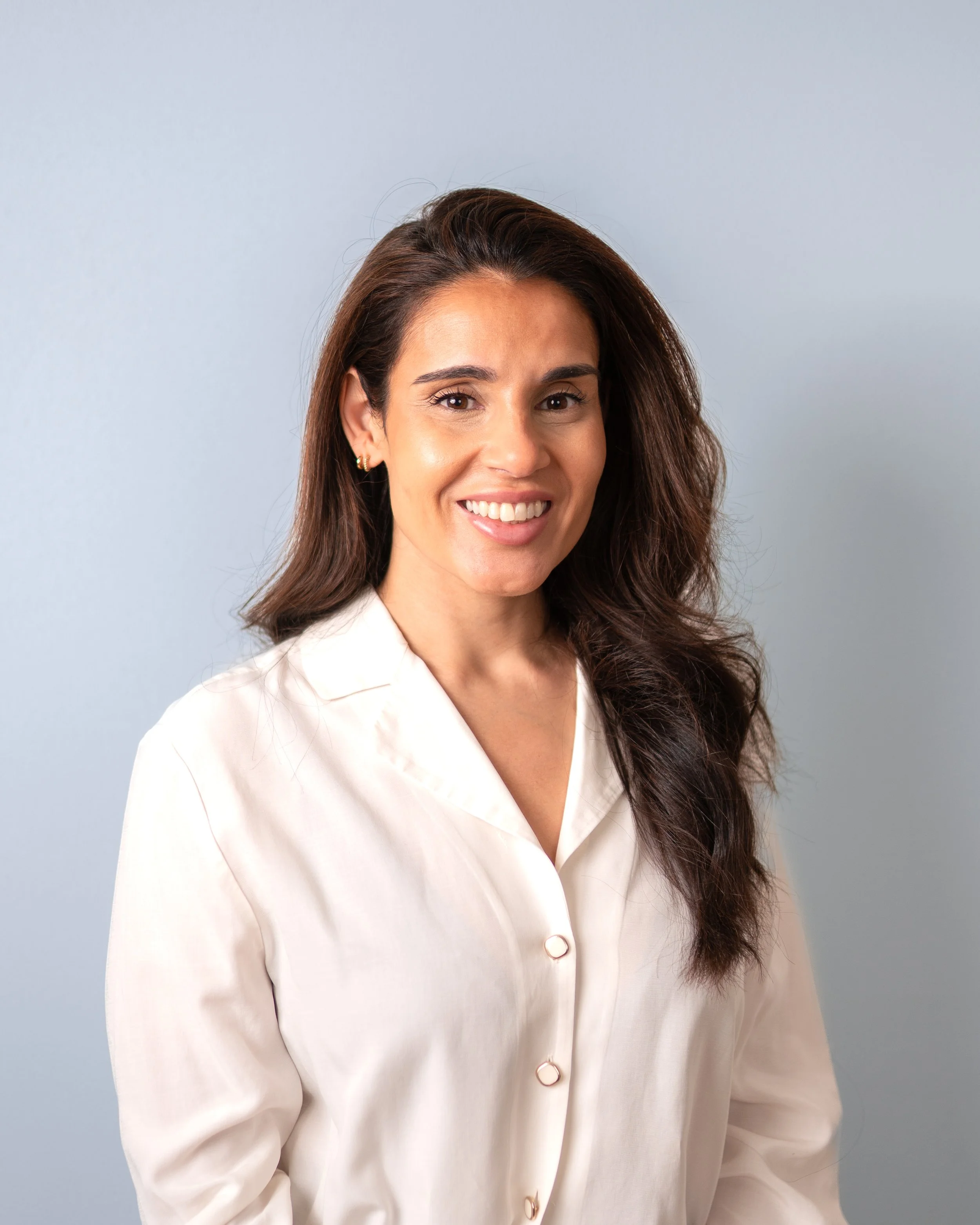 A woman with long, wavy brown hair, wearing a cream-colored blouse, smiling, standing against a light gray background.