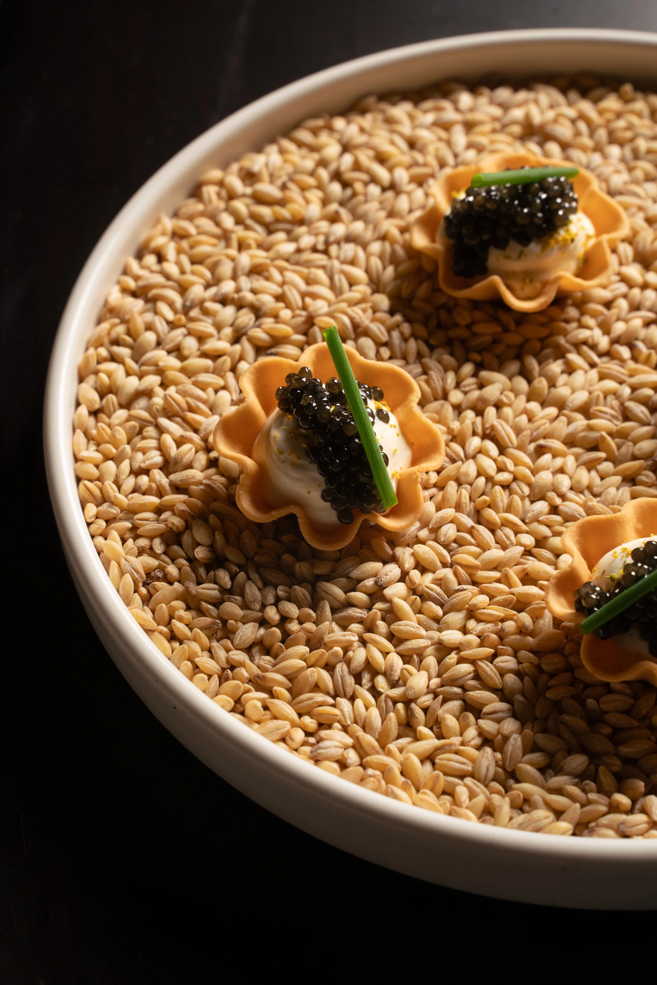 A white bowl filled with toasted sesame seeds, topped with small tart-like appetizers garnished with a dollop of white cream, black caviar, a green chive, and a green straw.