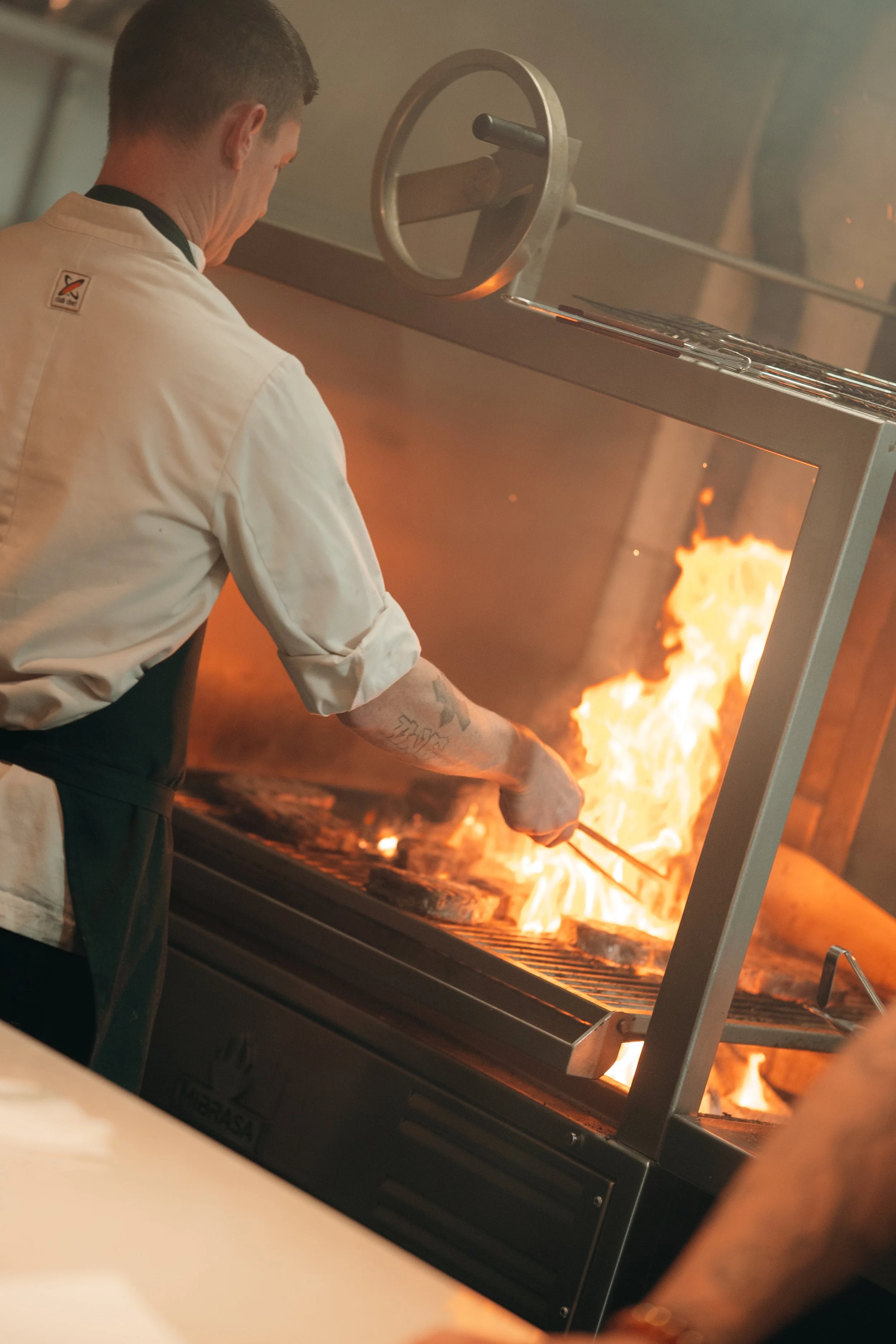 A chef grilling food on a large outdoor grill with flames.