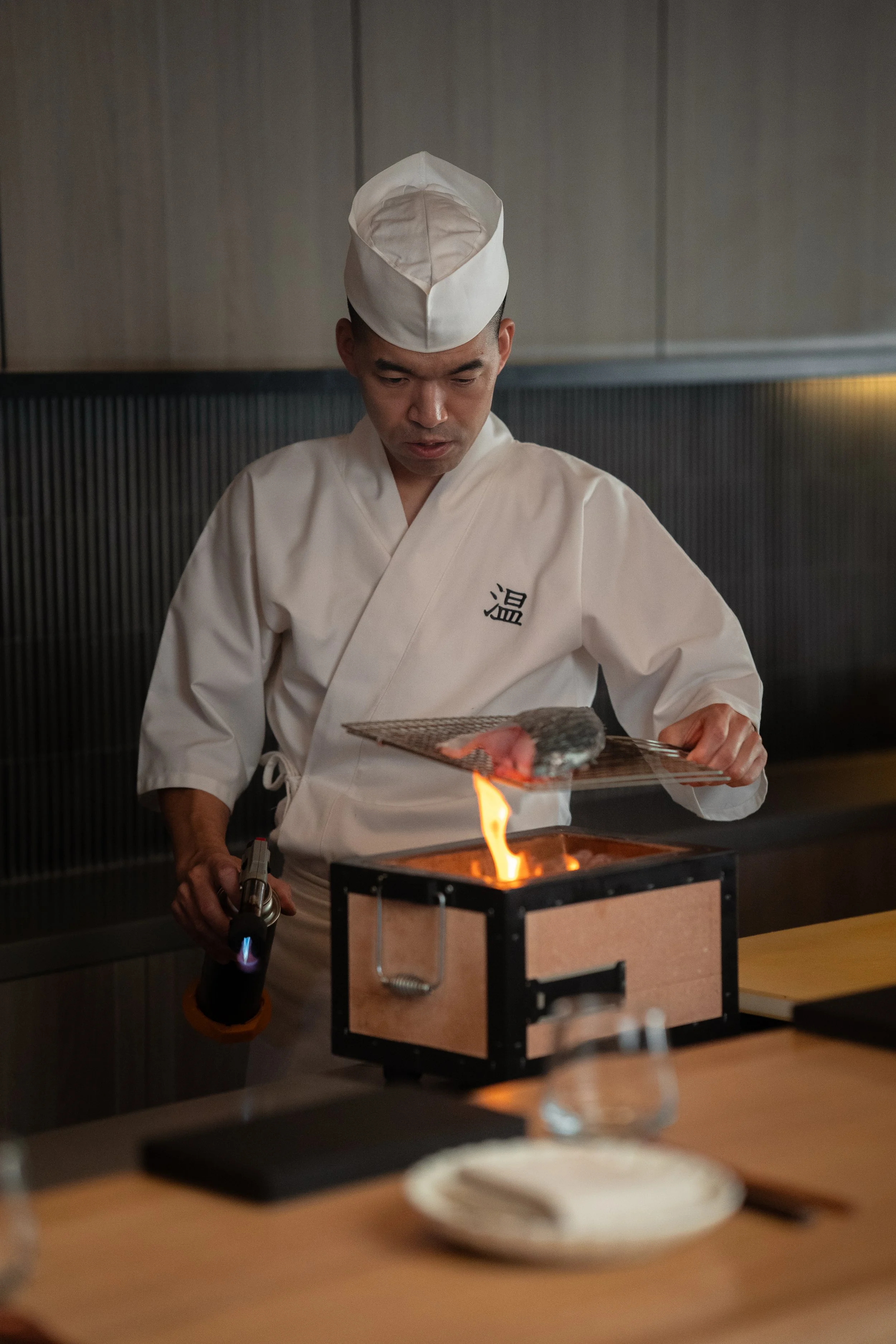 A chef in traditional Japanese attire is using a blowtorch to sear fish over a small flame on a grill in a modern restaurant setting.