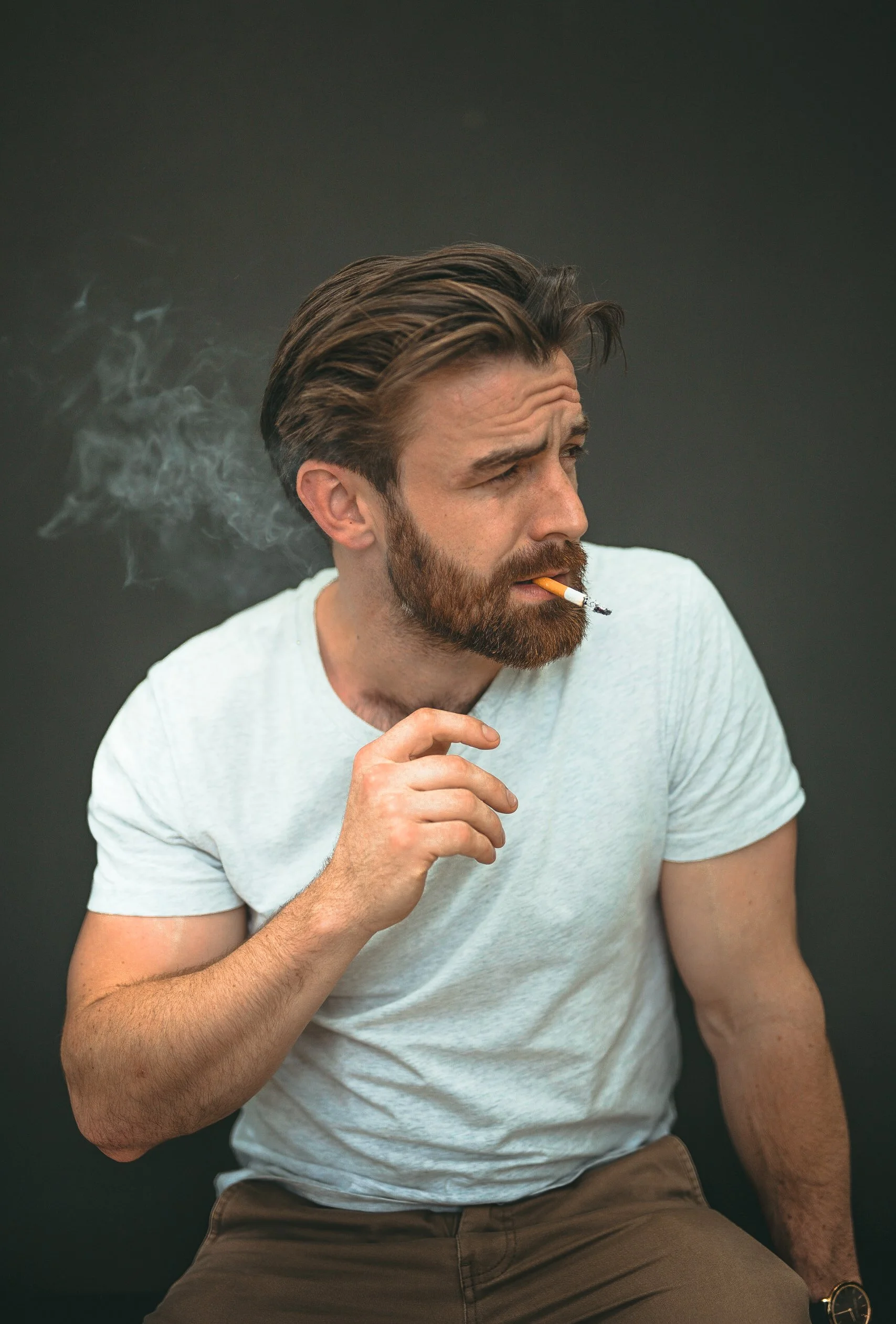 A man with brown hair and a beard, wearing a white t-shirt, smokes a cigarette against a dark background.