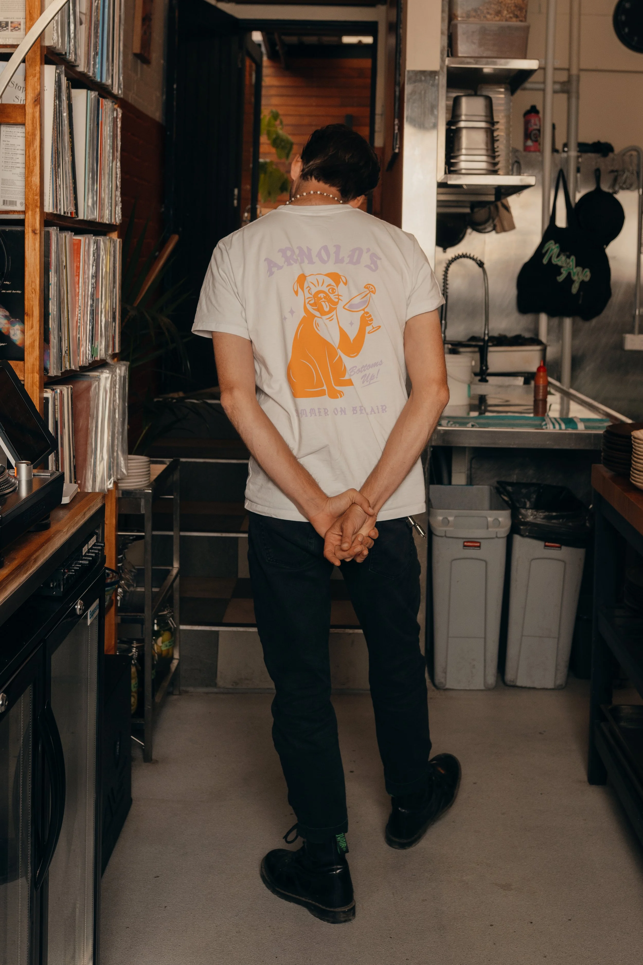 Person standing in a kitchen or cafe, wearing a white t-shirt with a cartoon tiger and the text "Arnold's Summer on Bear" on the back, with their hands clasped behind their back.