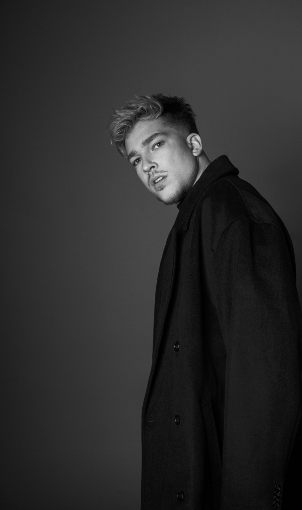 Black and white portrait of a young man with short, styled hair, wearing a dark coat, looking over his shoulder at the camera against a plain background.