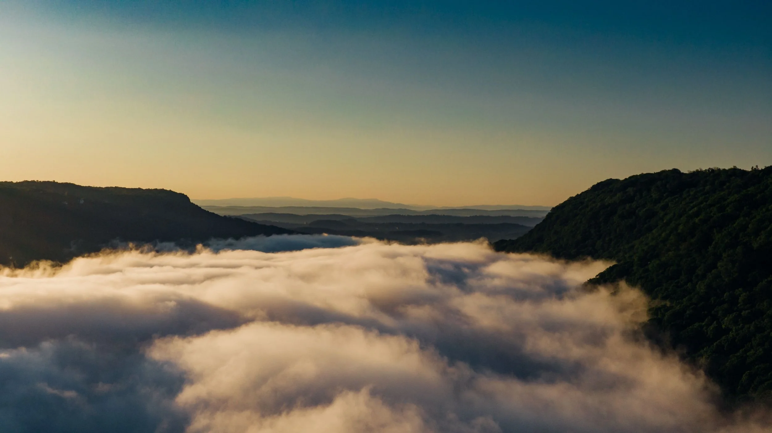 Aerial view of morning fog filling a Tennessee valley between forested ridgelines at sunrise, warm golden light above the clouds — Embodied Soul Psychotherapy®, telehealth across Tennessee
