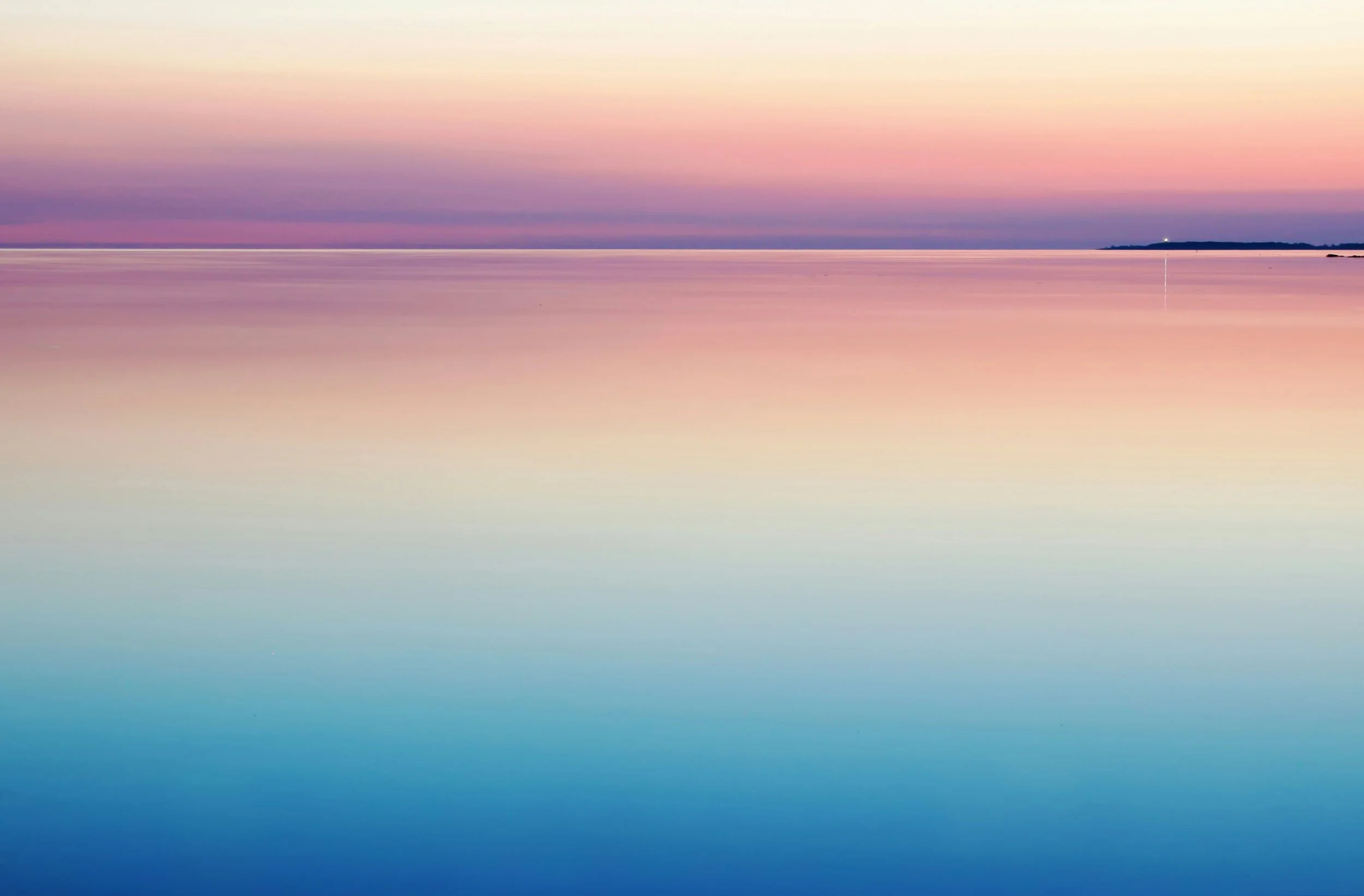 A calm body of water reflecting a pastel-colored sky at sunset or sunrise, with a distant horizon and a small light on the right.