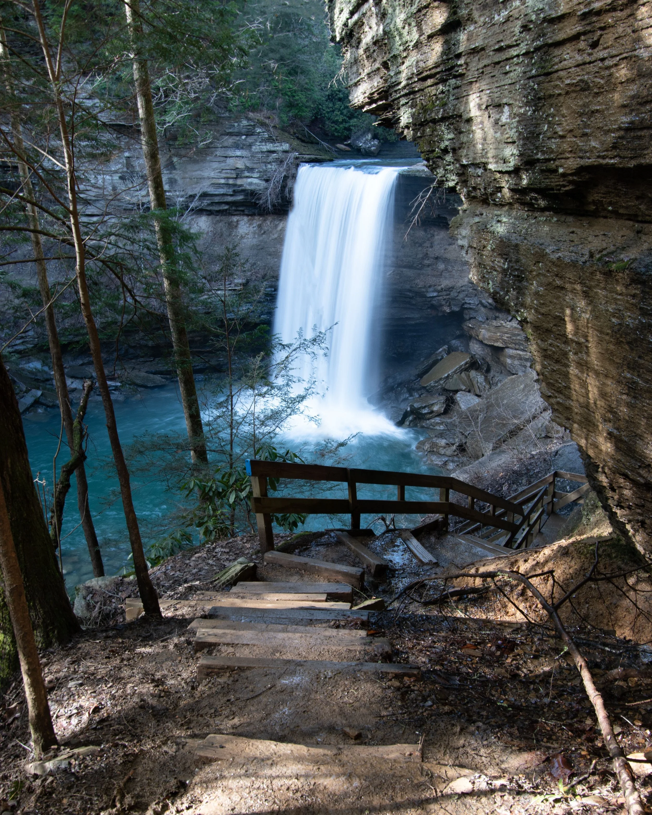 Wooden trail stairs descending toward a waterfall in a Tennessee sandstone gorge, misty blue-green pool below — depth-oriented psychotherapy, Tennessee telehealth
