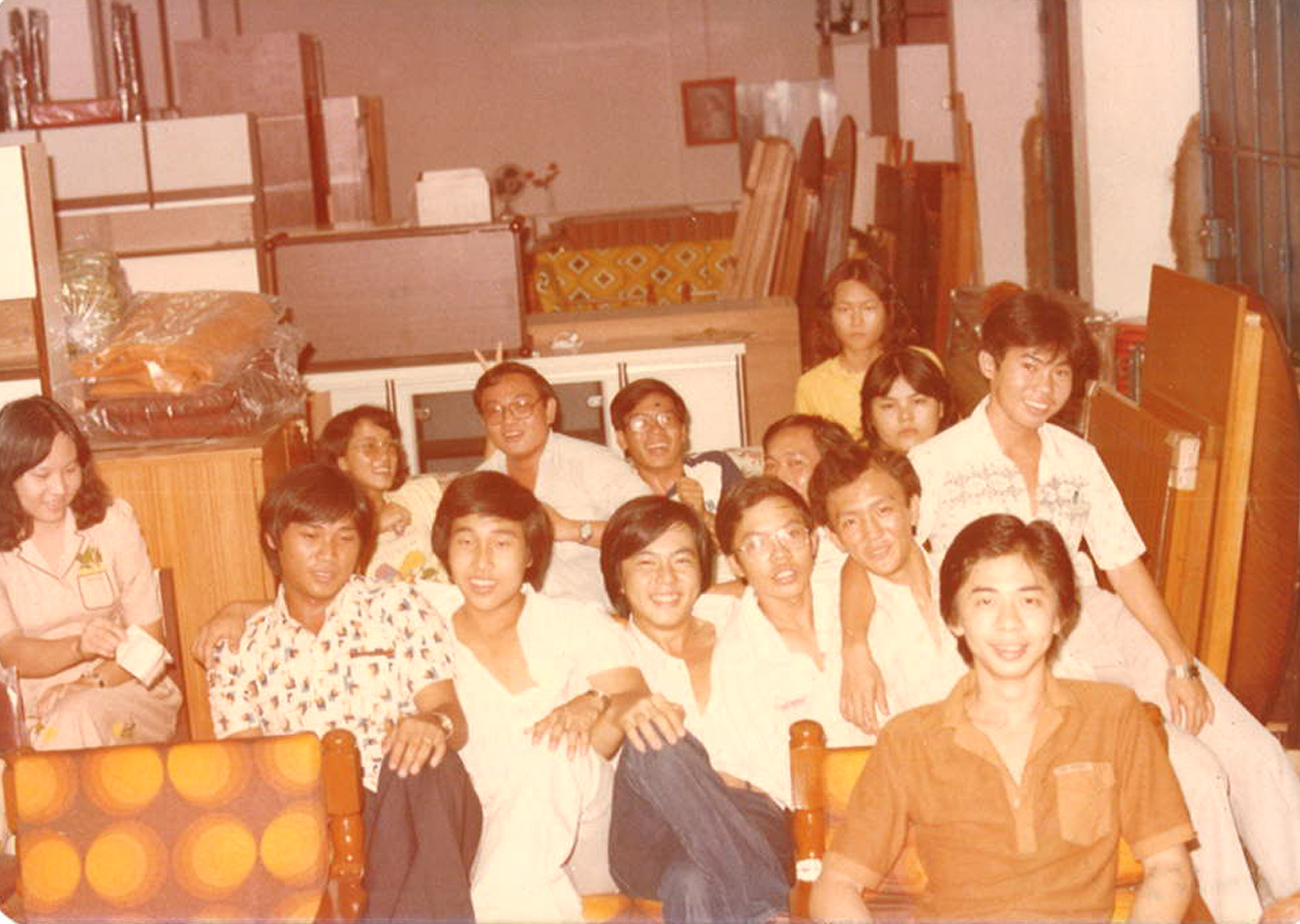 A group of people gathered together in a room, posing for a photo, with some seated and others standing behind them. The room has shelves and furniture, and the photo appears to be vintage, possibly from the 1980s or 1990s.