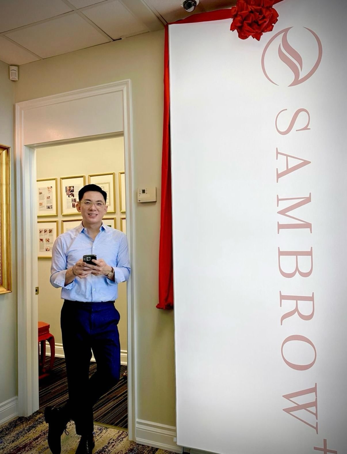 A man standing in a doorway with a smartphone in his hand, smiling. Behind him, framed pictures are visible on the wall. To the right, there is a large white sign with the text 'SAMBROW' and a logo at the top, decorated with a red ribbon and bow.